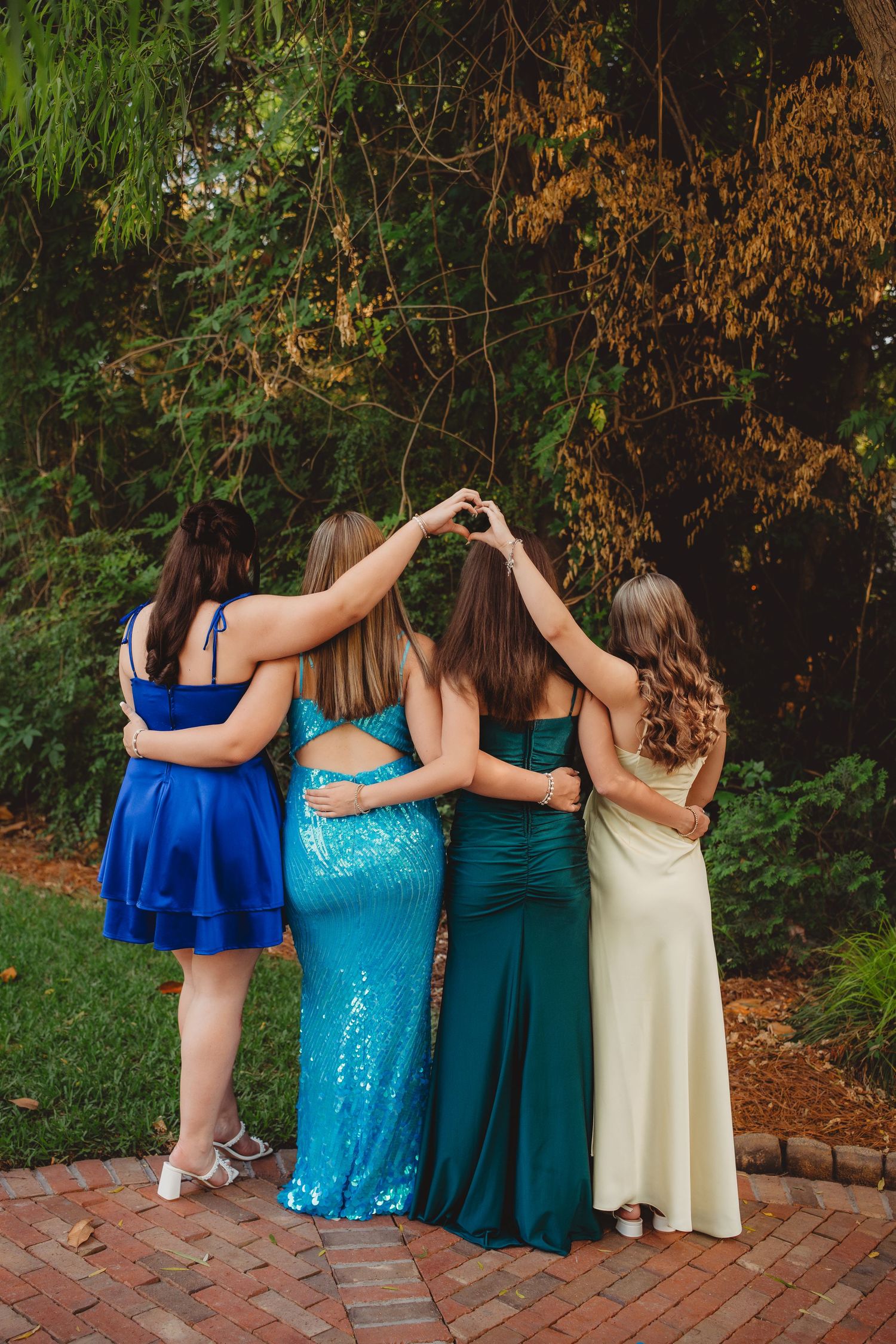 Four girls in colorful prom dresses making a heart shape with their hands while posing outdoors.