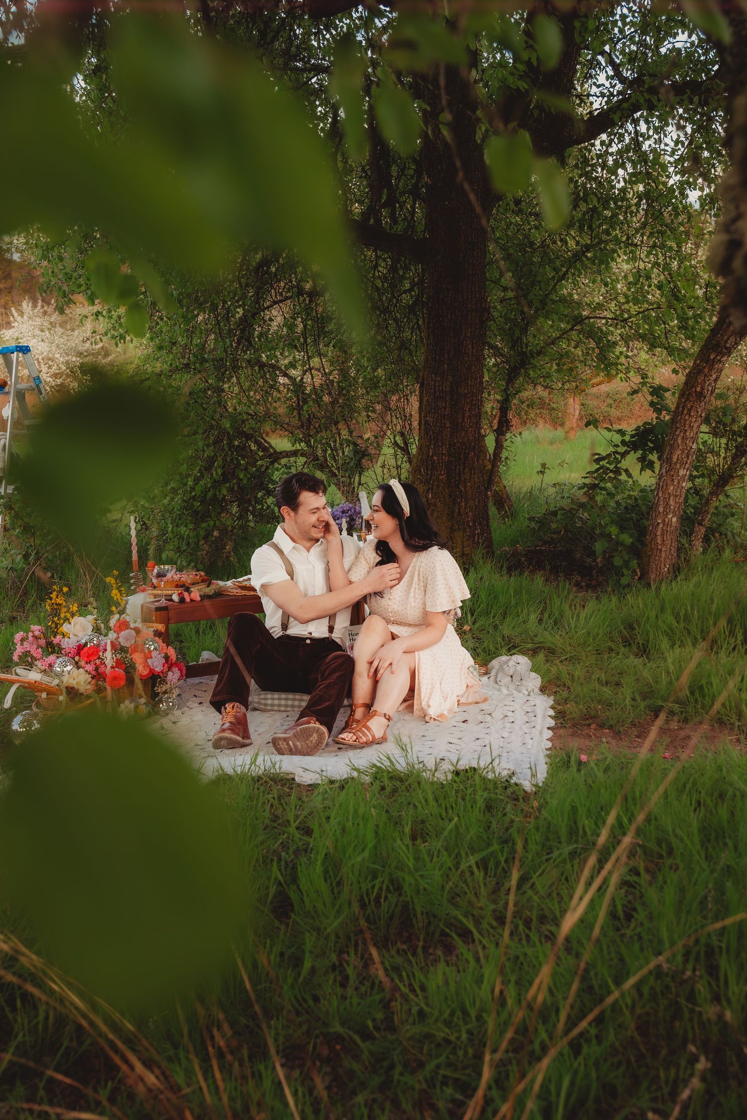 Couple sharing a romantic outdoor picnic on a blanket surrounded by lush greenery and colorful flowers.