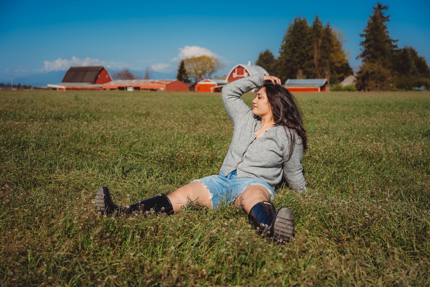 Young woman sitting in sunny green farm field with red barns in background.