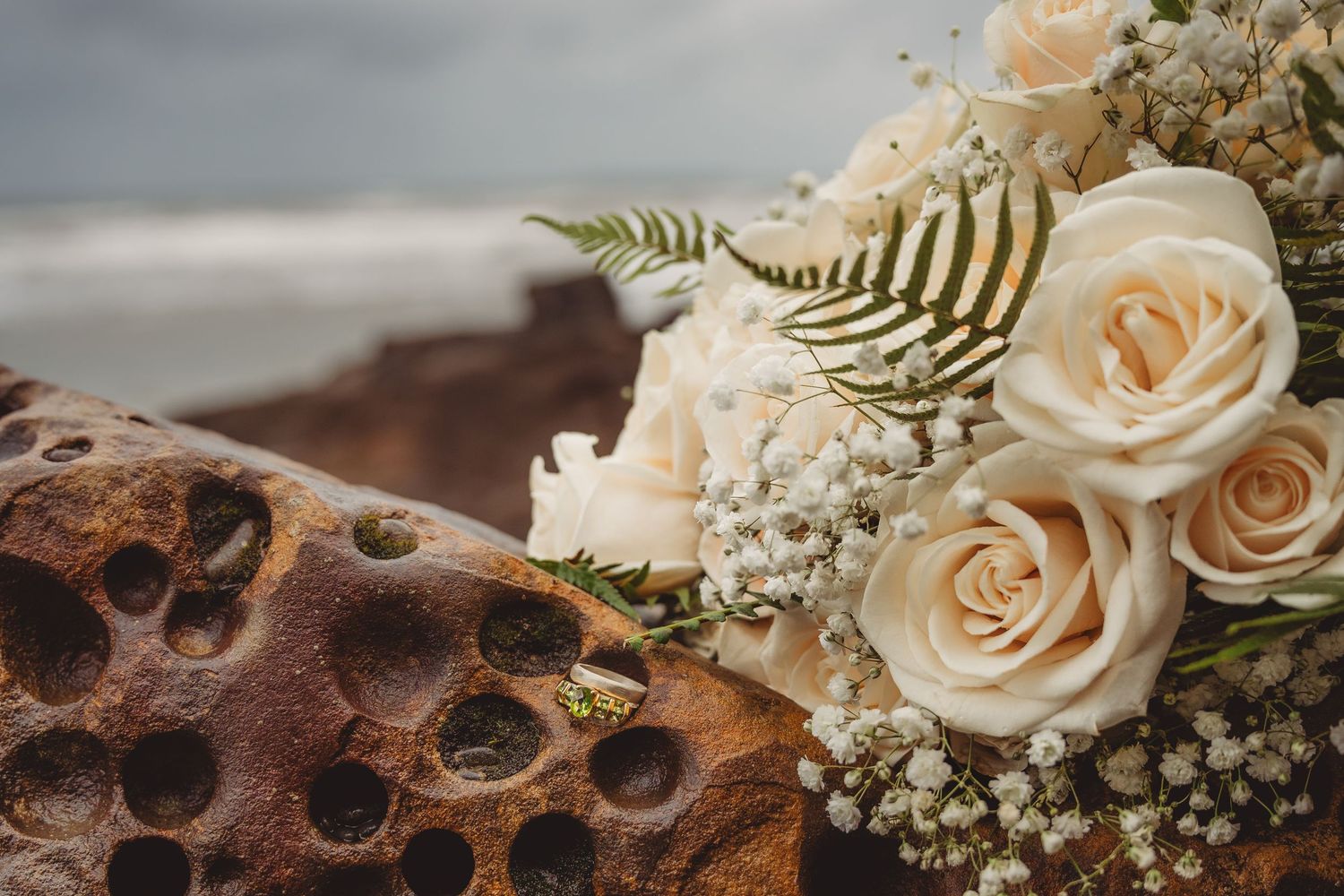 White roses and baby's breath bridal bouquet resting on textured rocks with ocean backdrop.