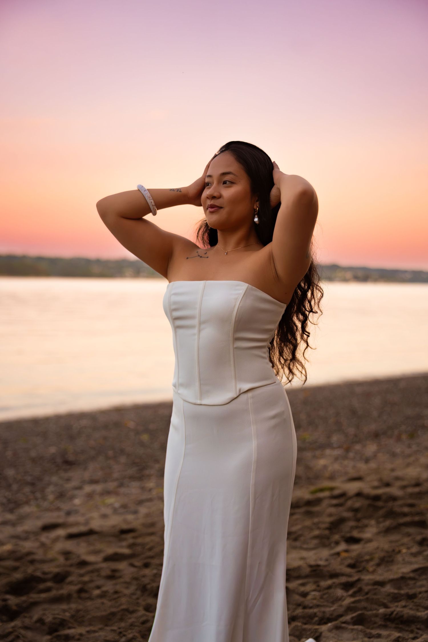 Woman in white strapless dress poses on beach at sunset with hands in hair.