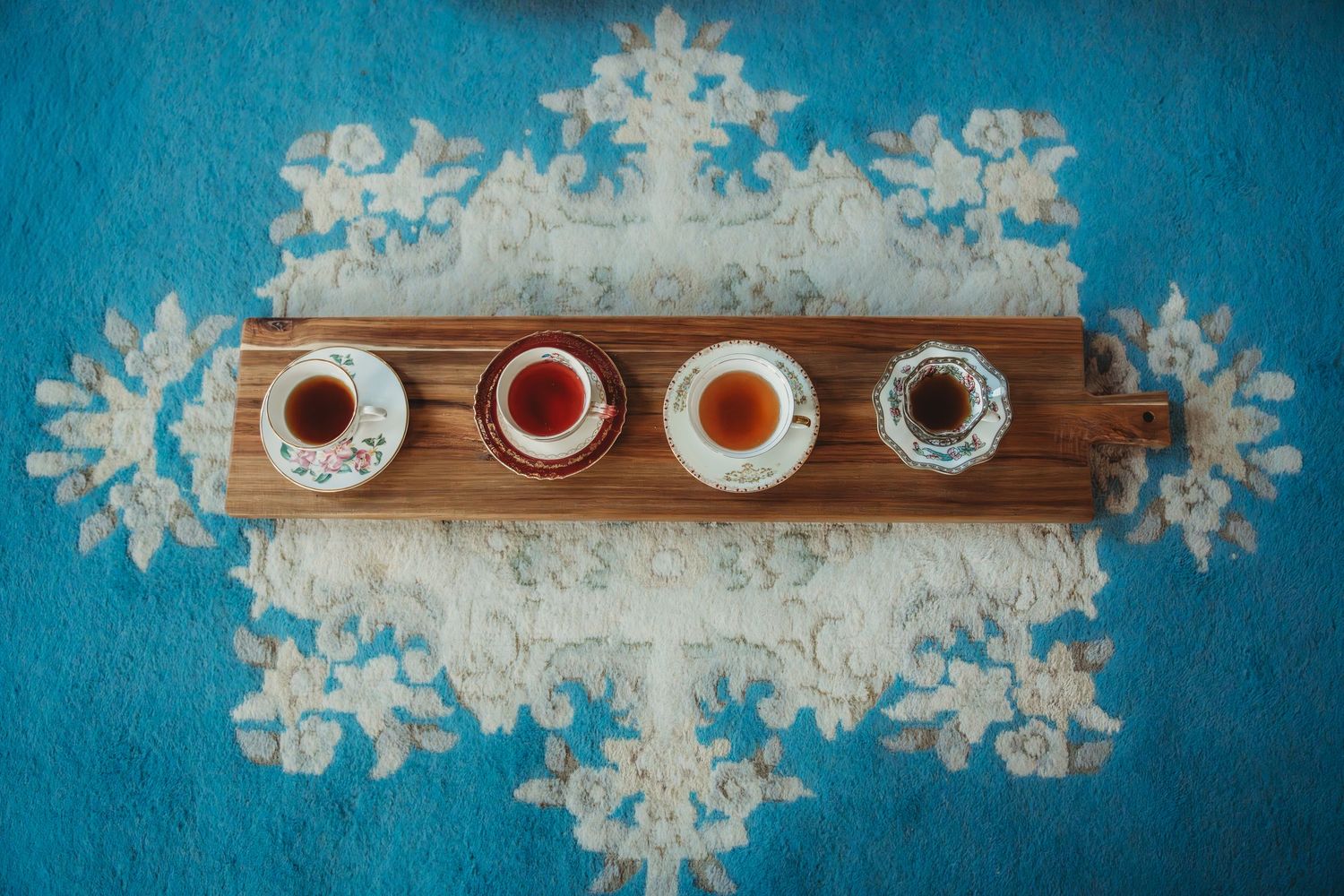 Four small cups of tea on a wooden tray atop a blue floral tablecloth, viewed from above.