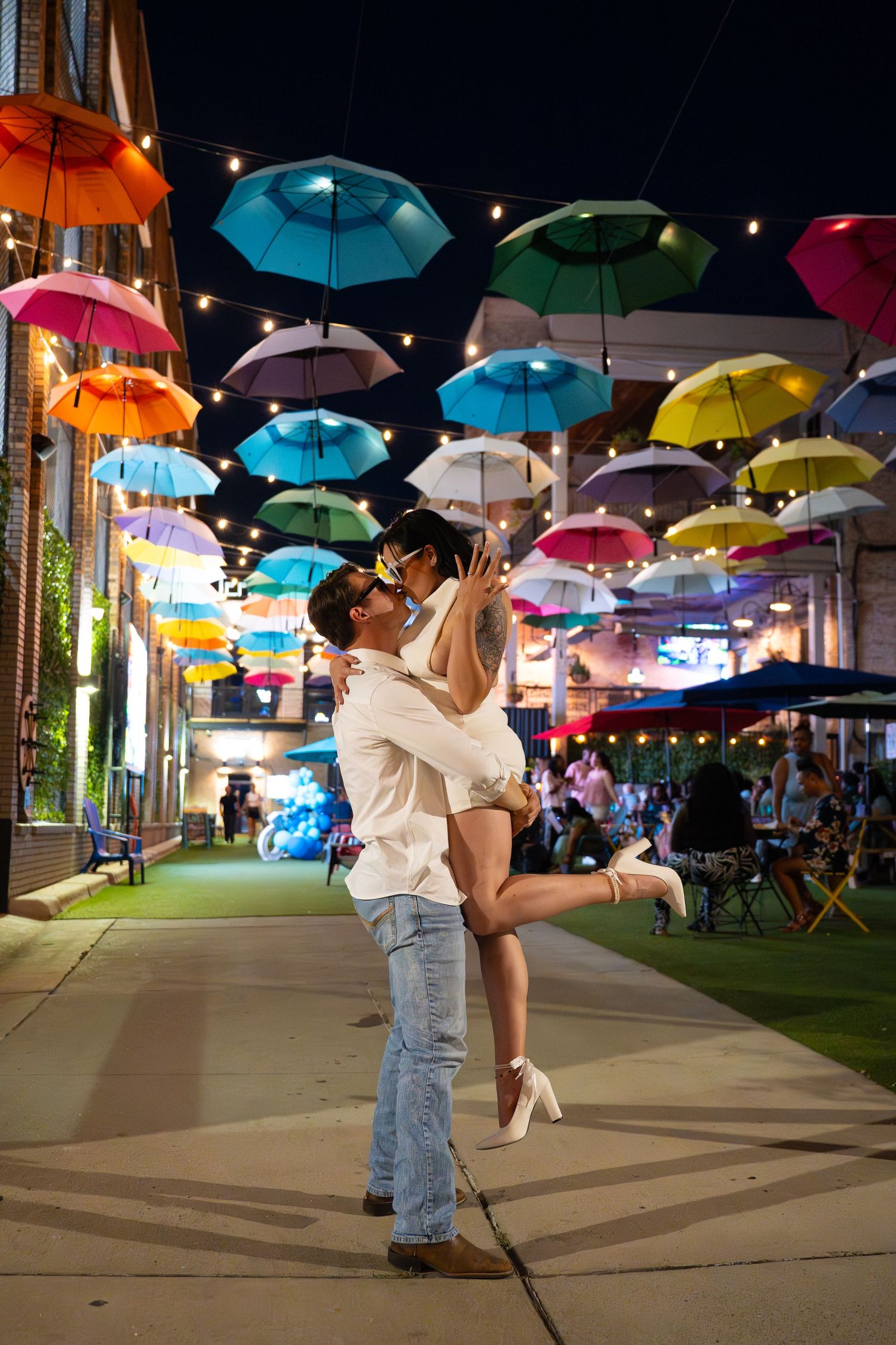 Couple embracing under a canopy of colorful floating umbrellas and string lights on a lively city street at night.