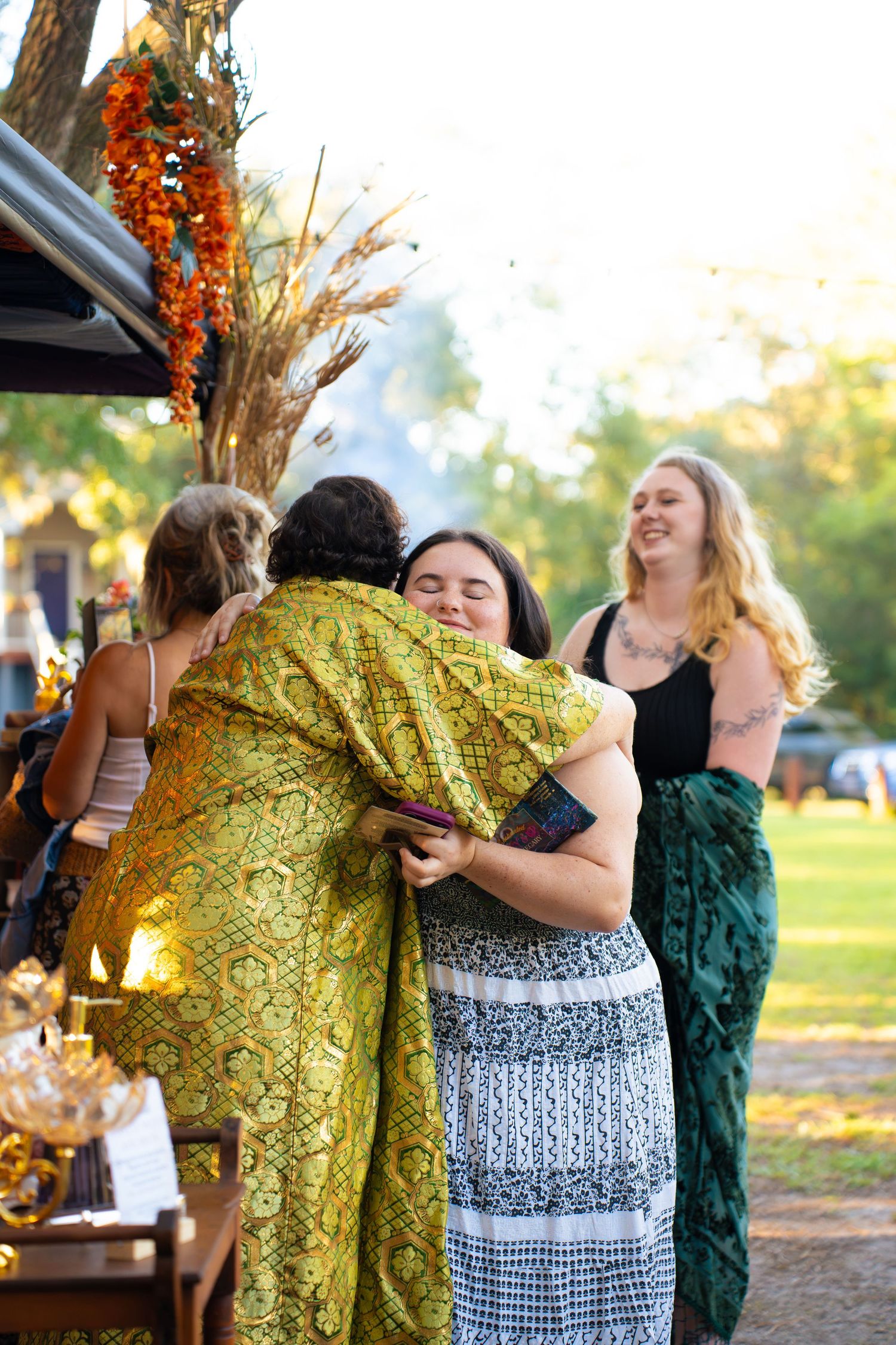 Two people in colorful patterned robes share a warm hug outdoors while smiling guests look on at a sunlit garden gathering.