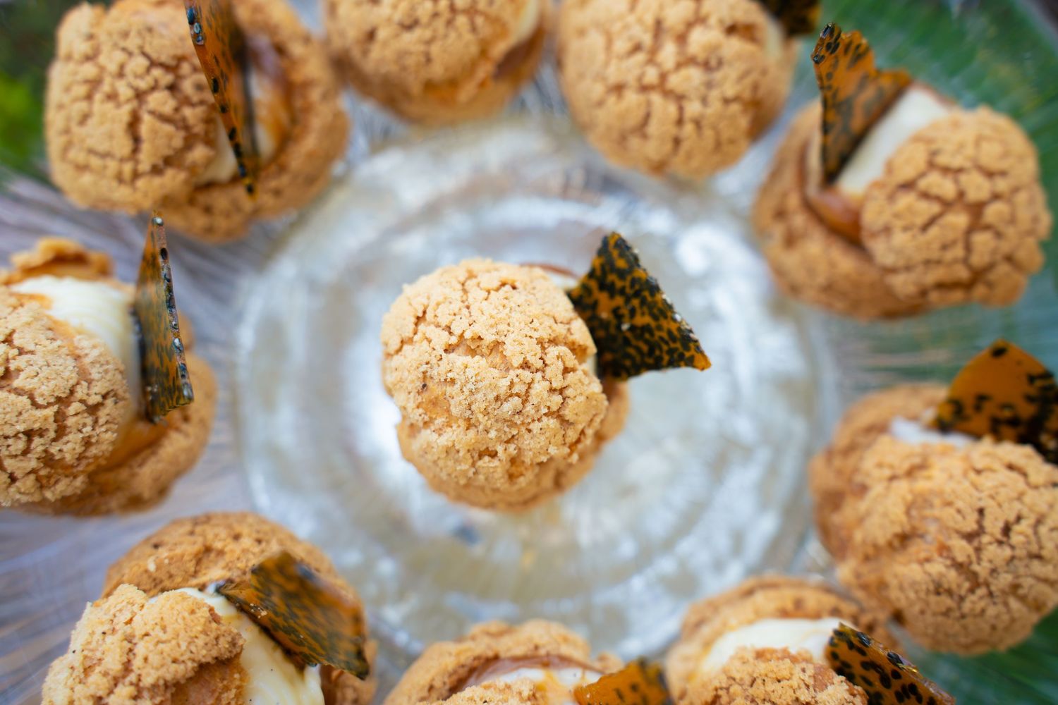 Overhead view of craquelin-topped choux pastry puffs filled with cream and garnished with caramelized shards on a clear glass plate.