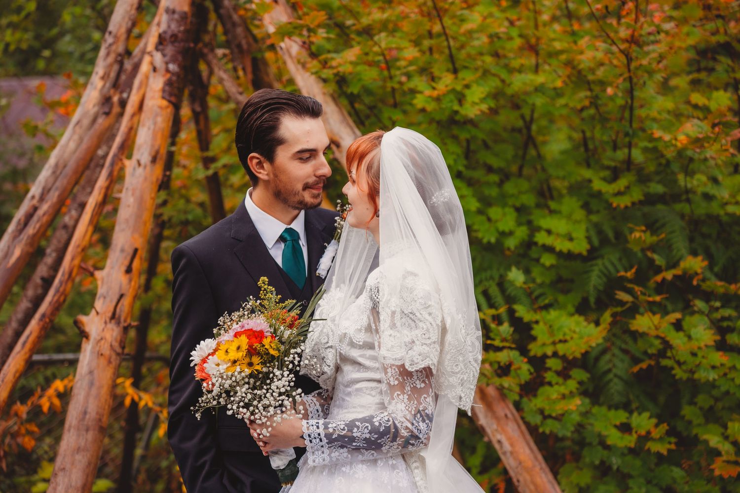 A bride and groom share a romantic moment outdoors in autumn, with colorful fall foliage and a rustic wooden structure behind them.