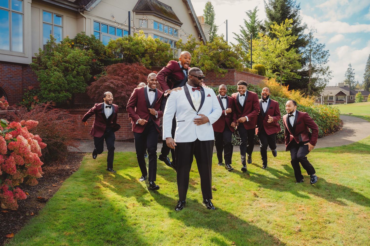 Groom in white tuxedo leads groomsmen in burgundy suits running on green lawn outside elegant venue.