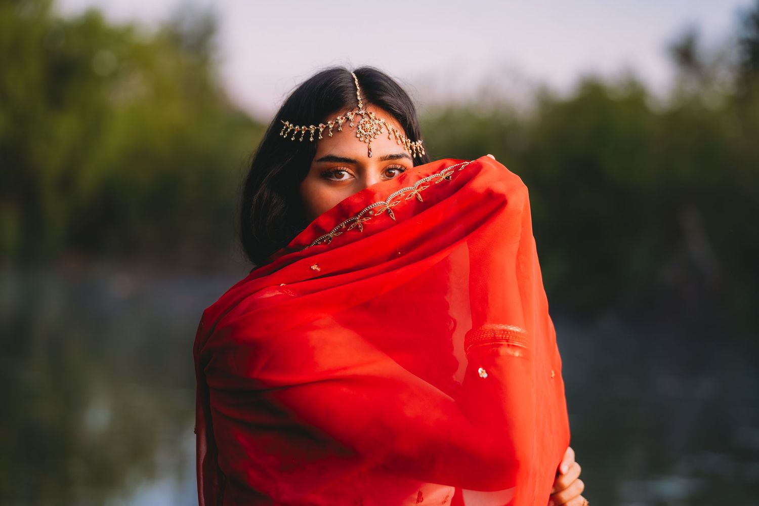Indian woman in red saree covering face with dupatta near a serene waterway.