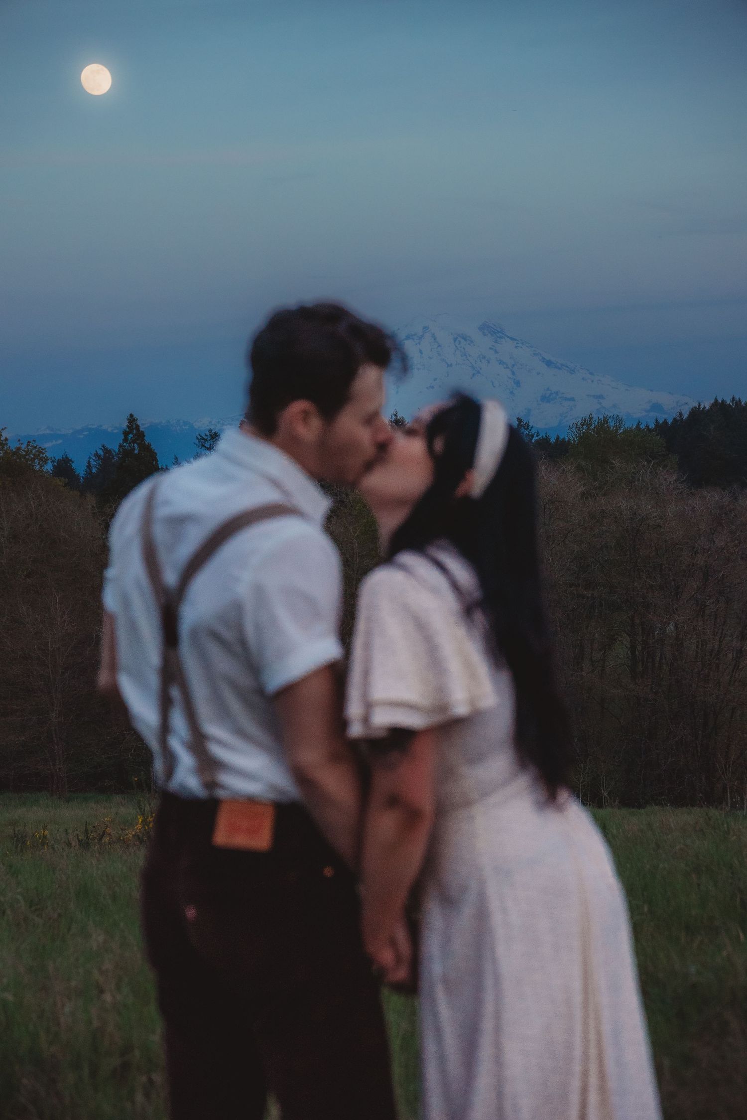 Couple sharing a romantic kiss at dusk with a full moon and mountain backdrop in a scenic outdoor setting.