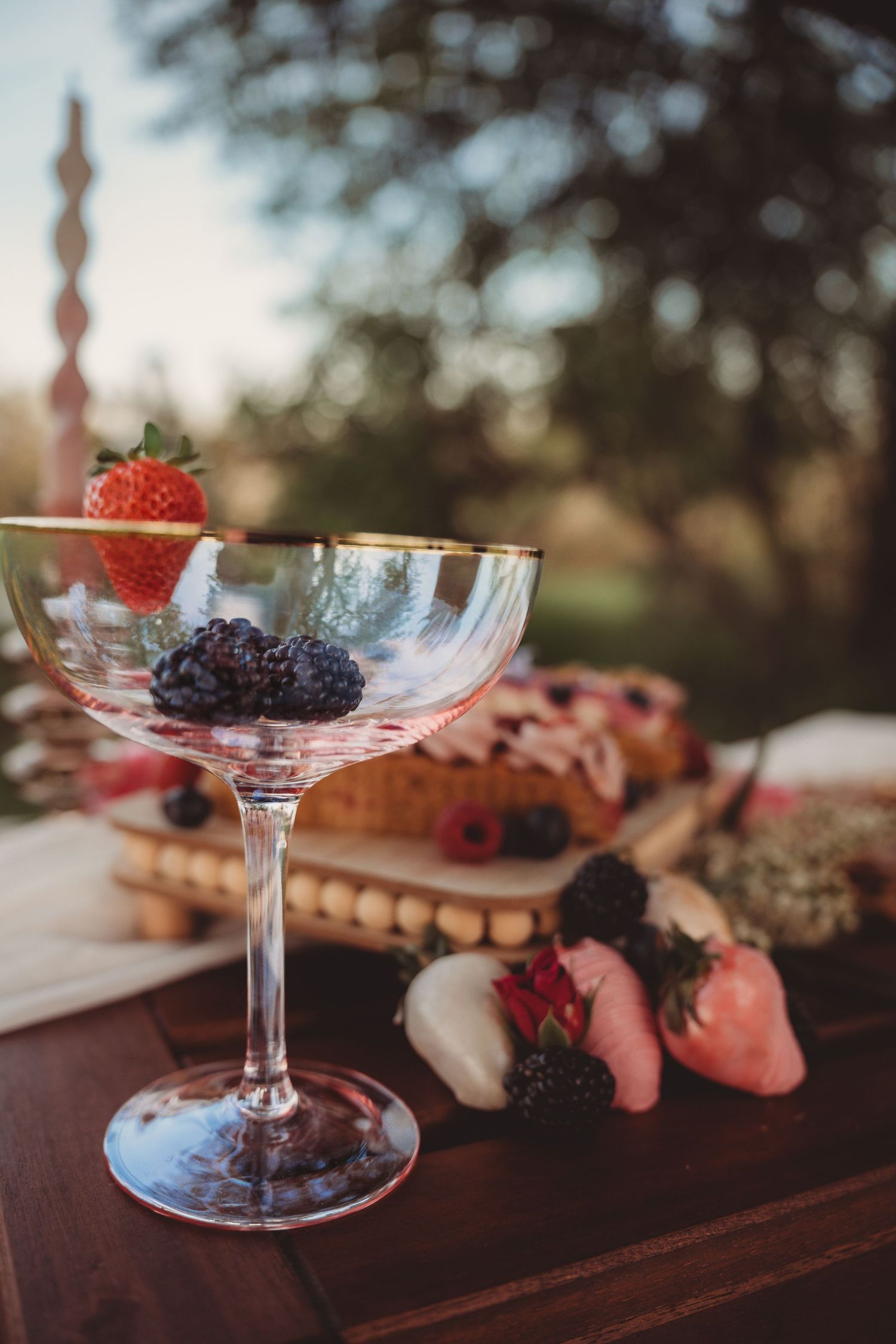 Elegant coupe glass with fresh strawberries and blackberries on a wooden table with a dessert charcuterie board outdoors.