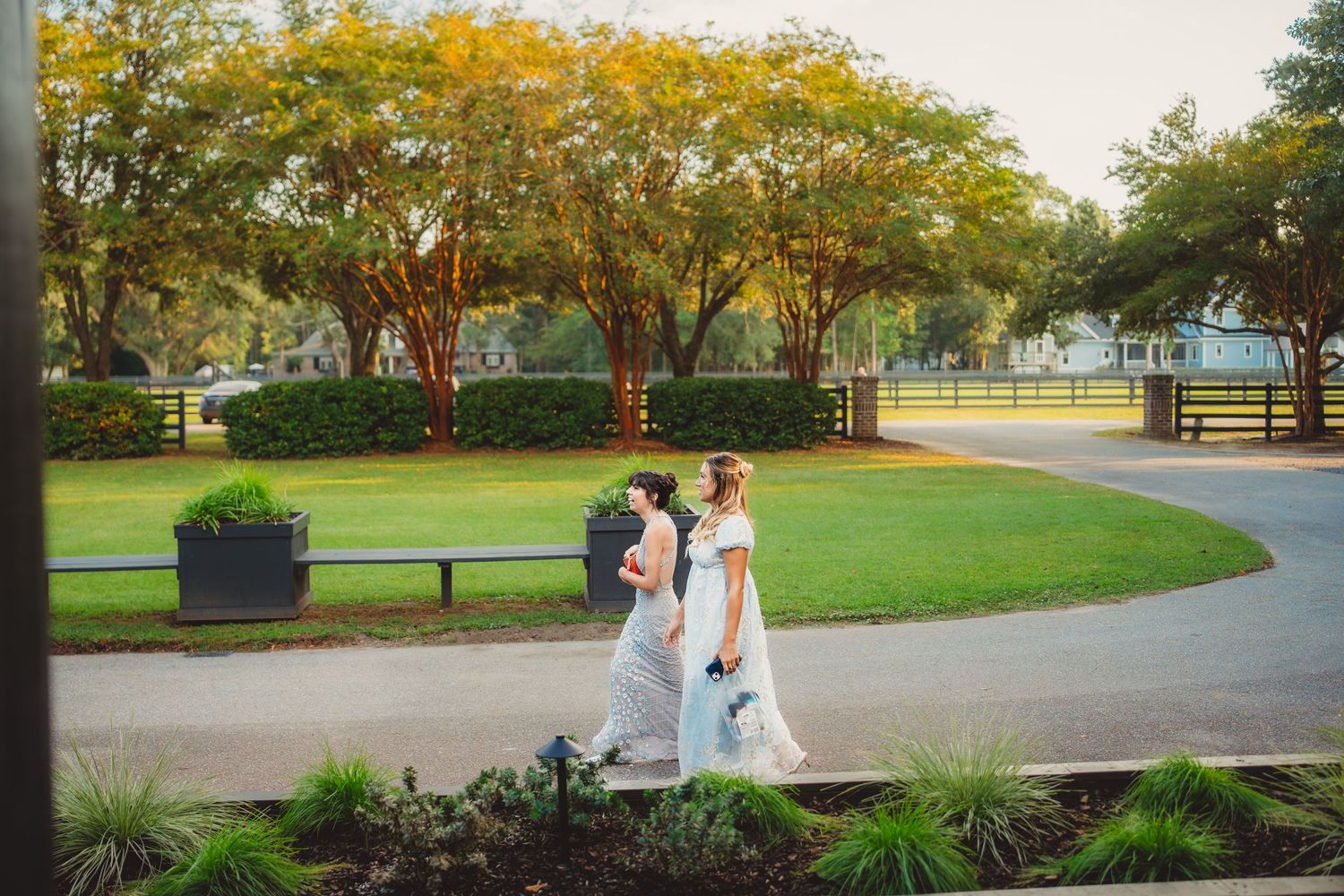 Two brides in white gowns walk together along a curved path surrounded by lush green trees at an outdoor wedding venue.