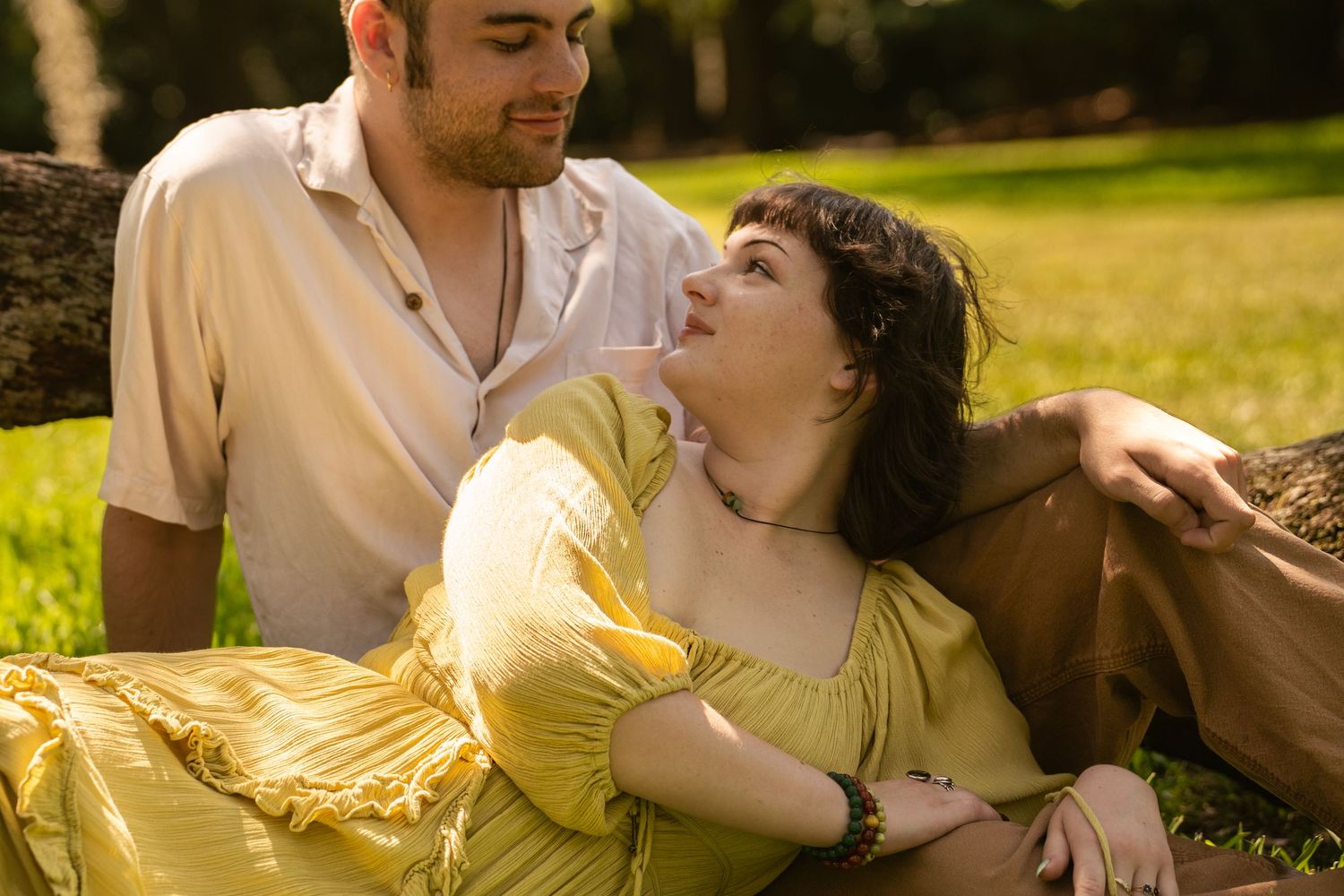 A couple relaxes on grass in a sunny park, woman in yellow dress gazing up at man leaning against a tree.
