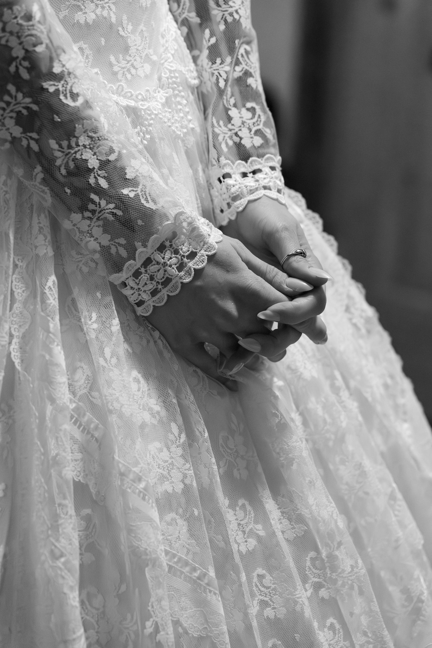 Black and white close-up of a bride's clasped hands in lace wedding dress sleeves with a delicate ring.