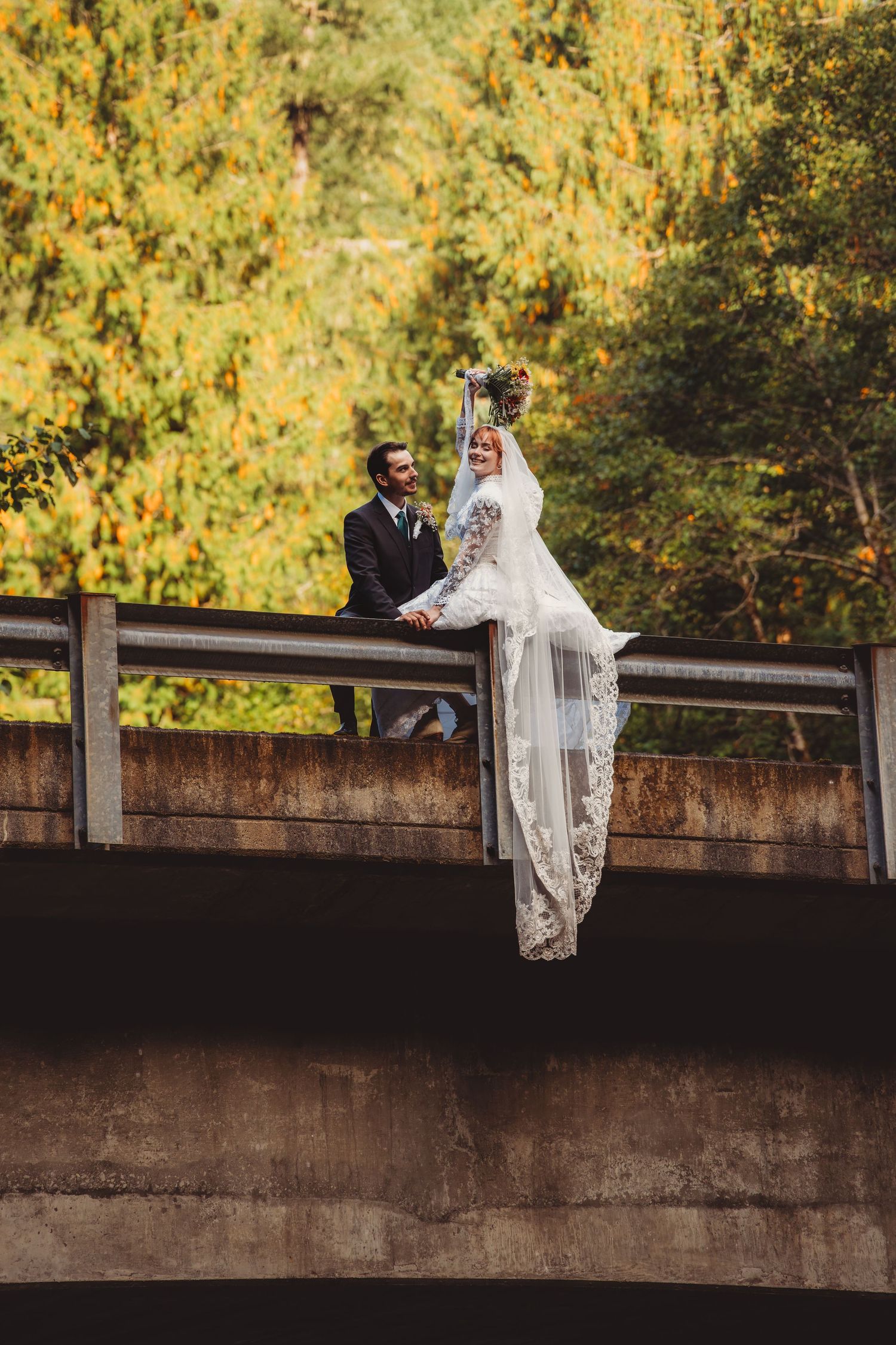 Bride in lace gown and veil sits on bridge railing with groom in black suit, autumn trees in background.