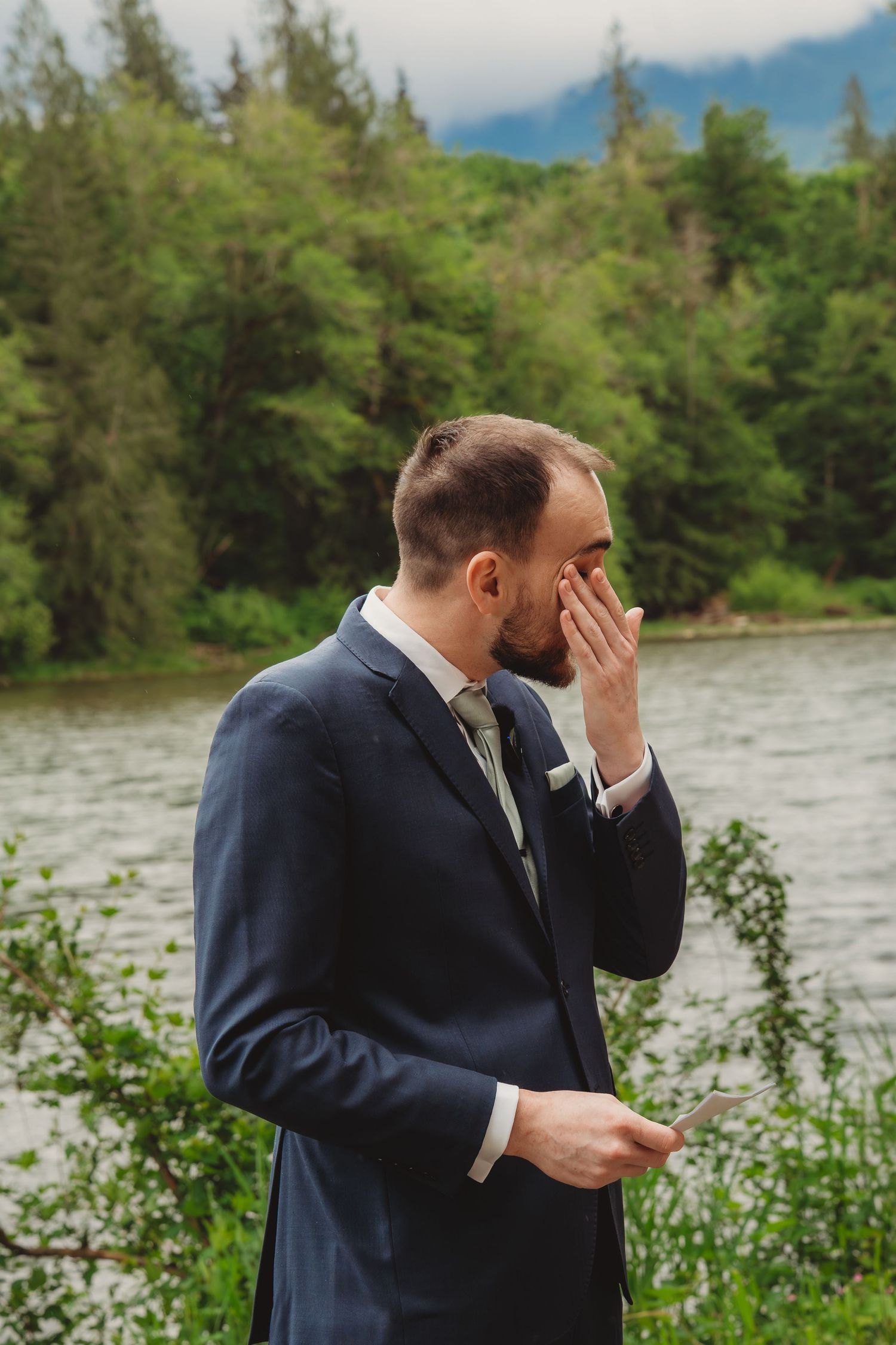 Emotional groom in navy suit wiping tears by a scenic lake and forest backdrop on his wedding day.