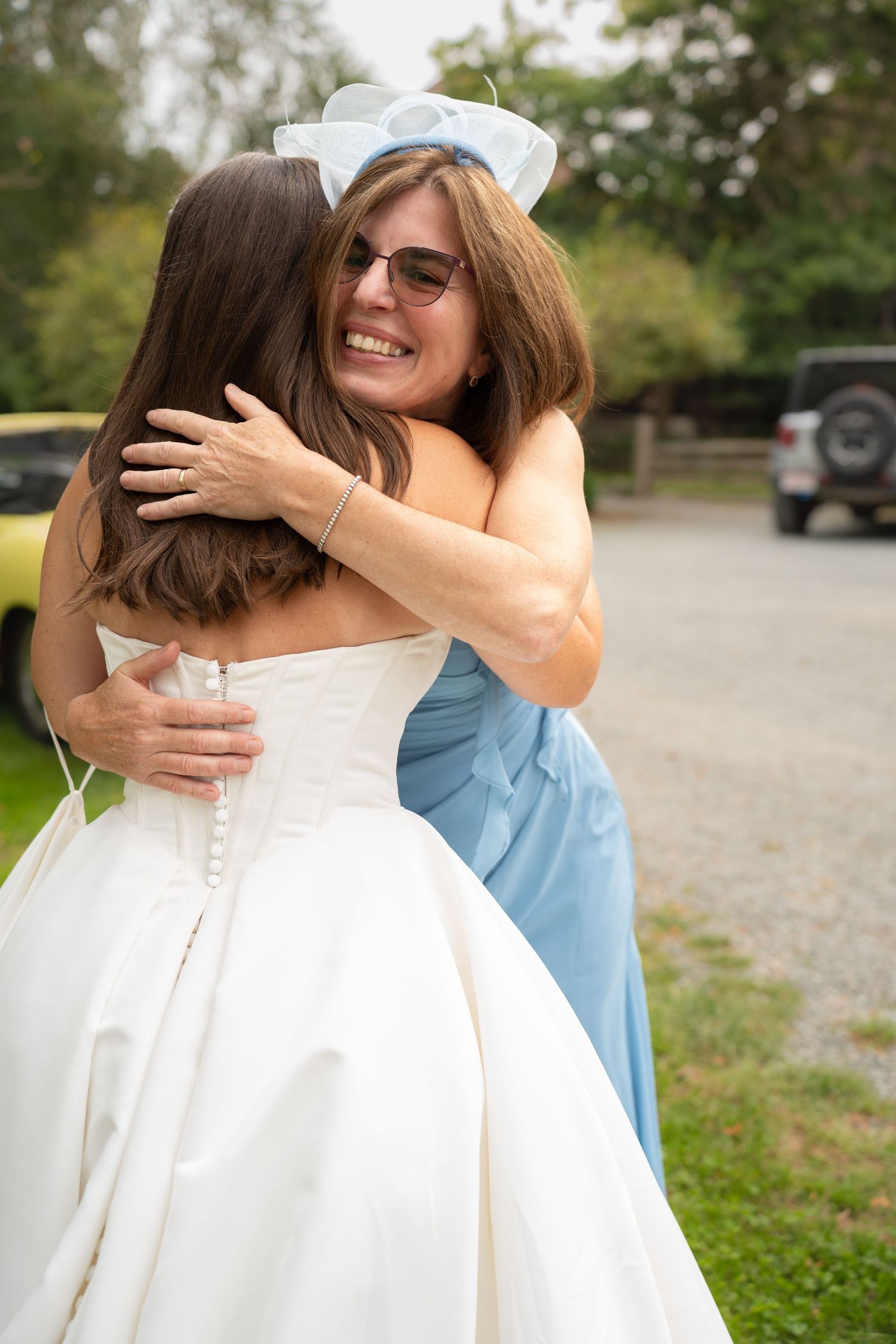 Bride in white gown hugging woman in blue bridesmaid dress with bow headband at outdoor wedding celebration.