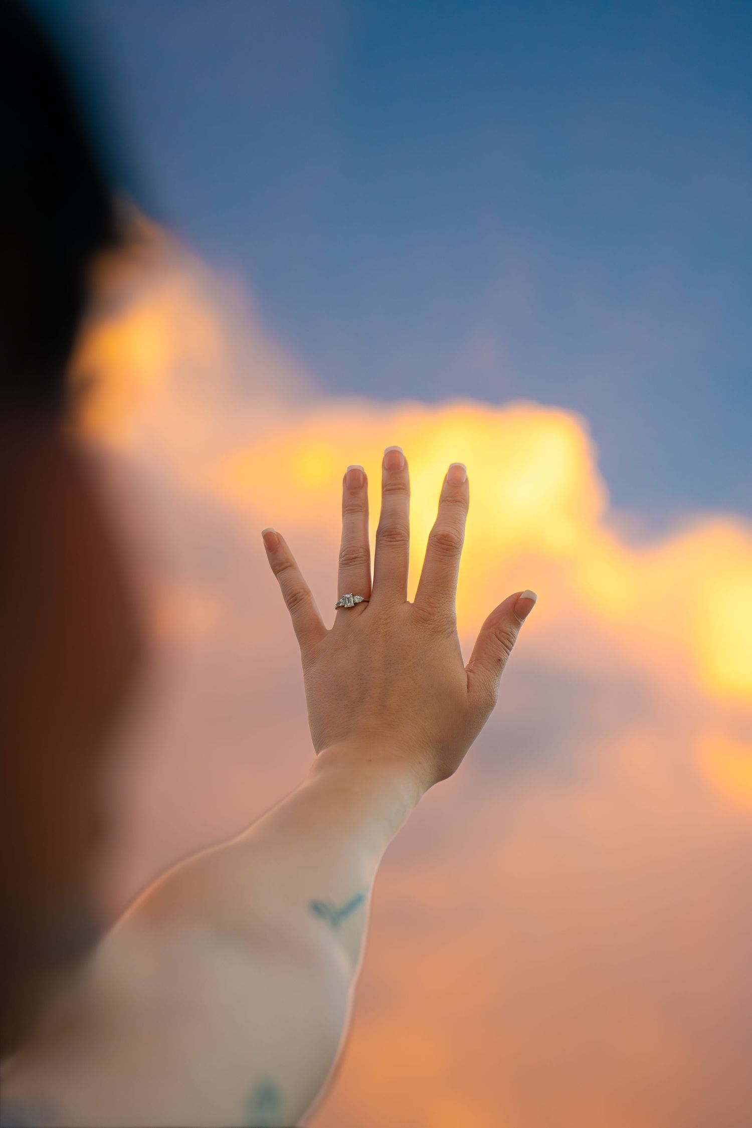 Outstretched hand wearing a ring and a small bird tattoo on the forearm, reaching toward glowing orange clouds in a blue sunset sky.