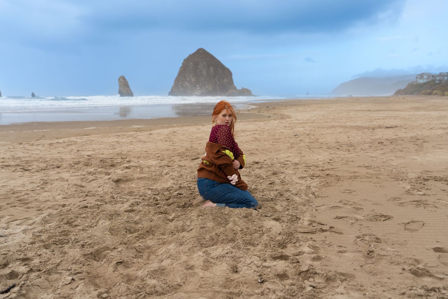 A child kneels on sandy Cannon Beach with iconic Haystack Rock in the background under a cloudy sky.