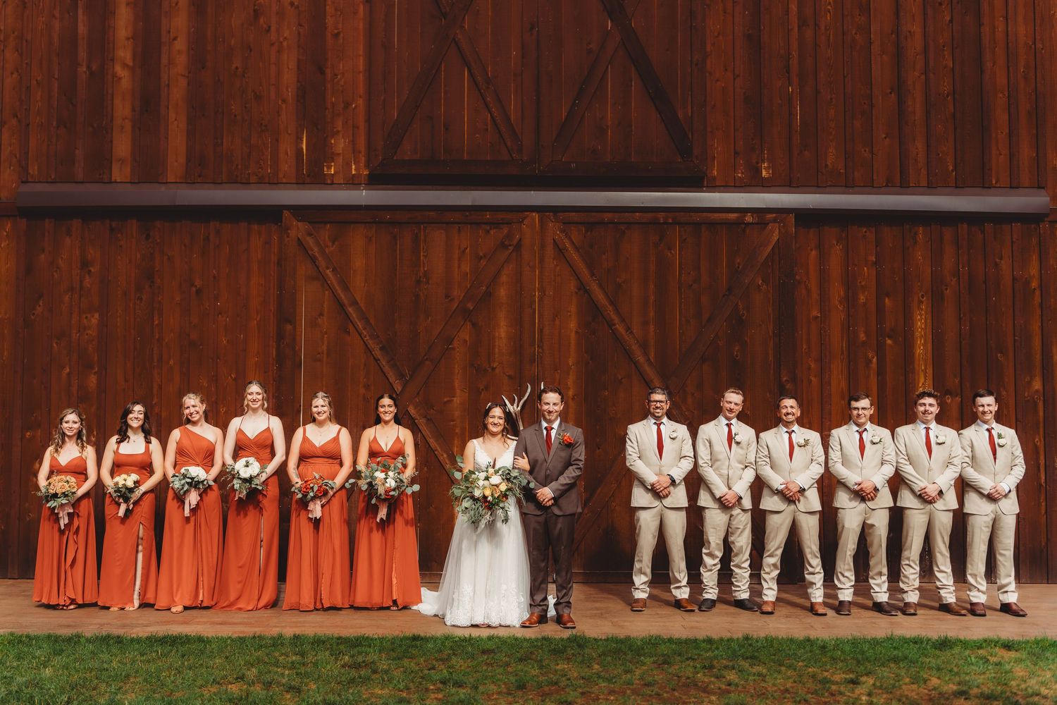Wedding party poses before a rustic red barn, bridesmaids in orange gowns and groomsmen in tan suits flanking the couple.