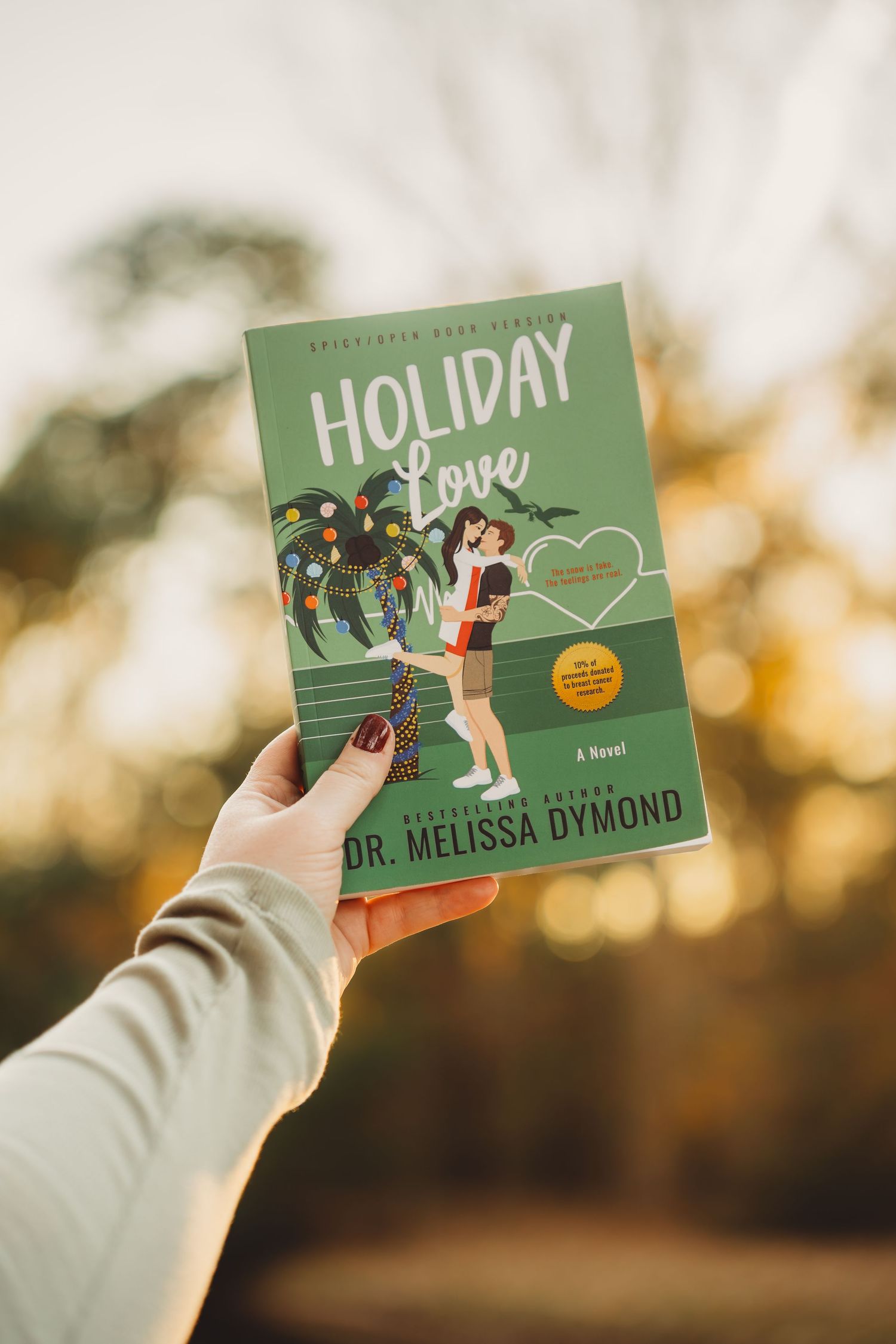 Hand holding Holiday Love novel by Dr. Melissa Dymond with festive cover art, photographed outdoors with bokeh background.