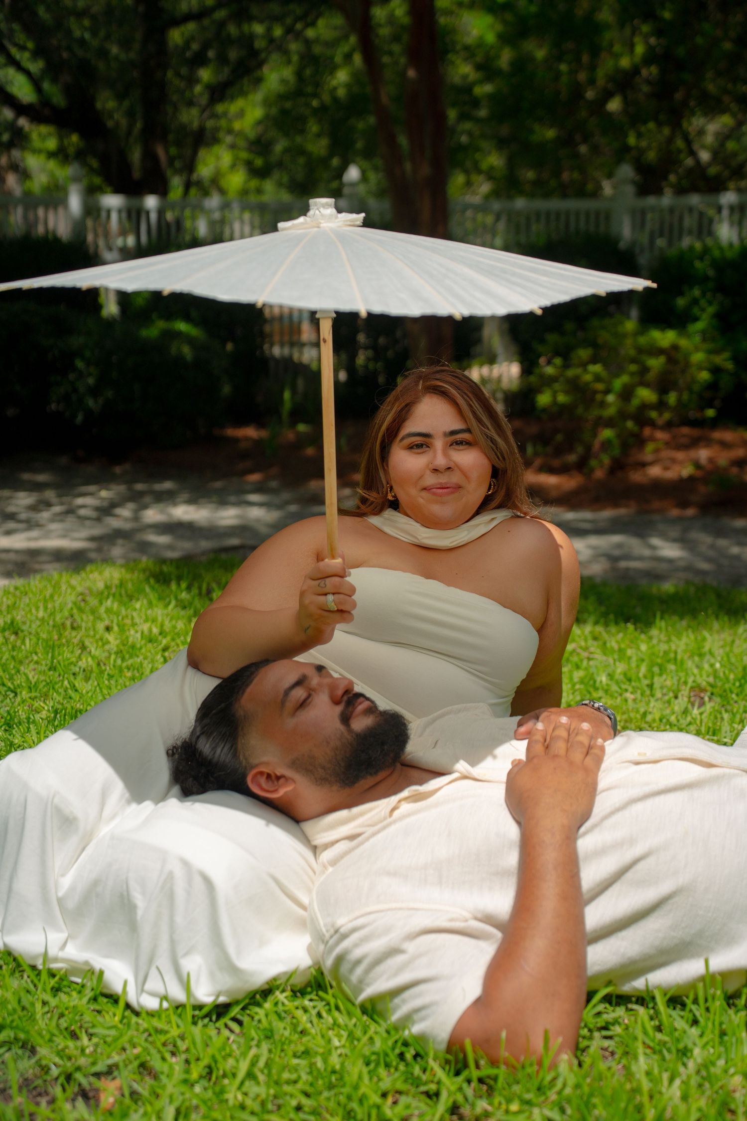 A woman in white holds a parasol over a bearded man lying on grass in a lush outdoor setting.