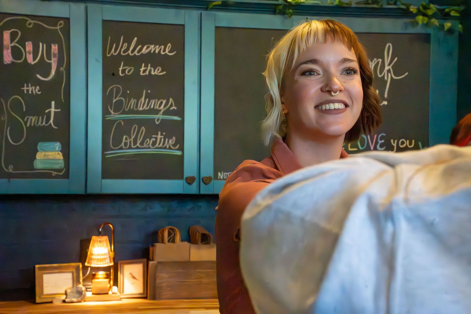 Smiling blonde woman behind teal bookstore counter with Welcome to the Endings Collective chalkboard sign.