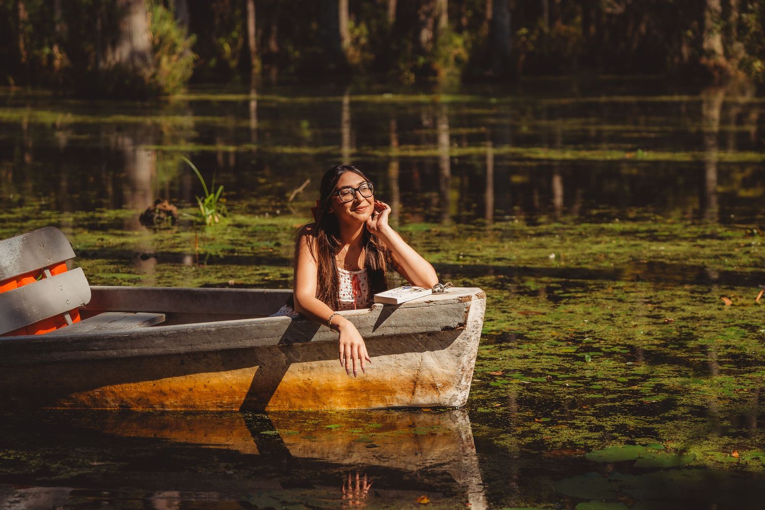 Woman relaxing in a wooden rowboat on a serene, green algae-covered lake surrounded by cypress trees. cypress gardens