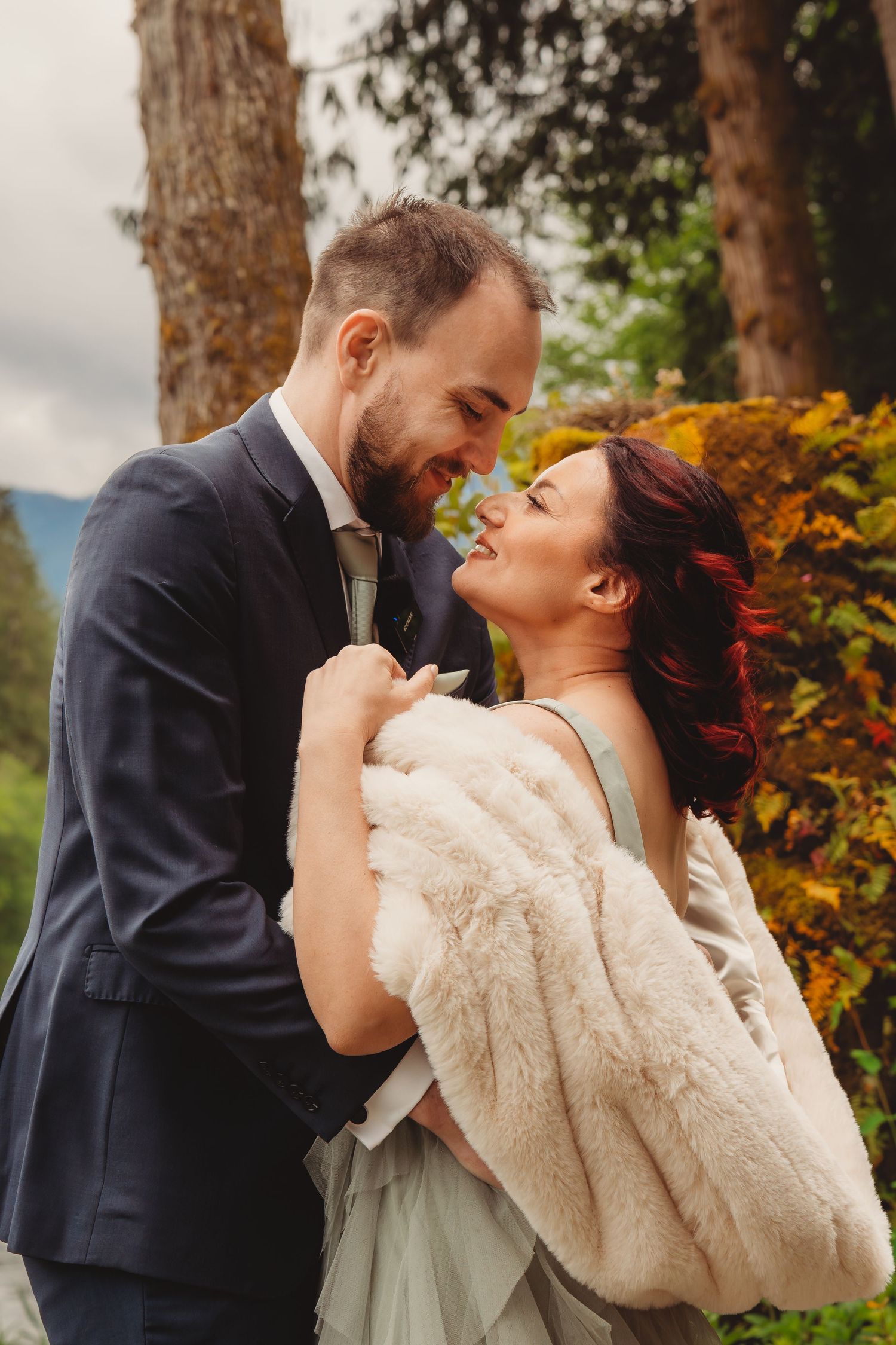 A bride in a fur wrap and groom in a navy suit share a romantic moment outdoors among autumn foliage.