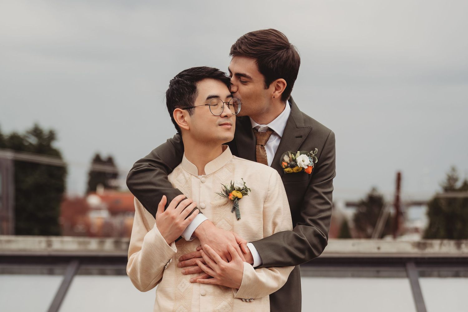 Two grooms share a tender moment, one kissing the other on the cheek, both wearing boutonnieres outdoors.