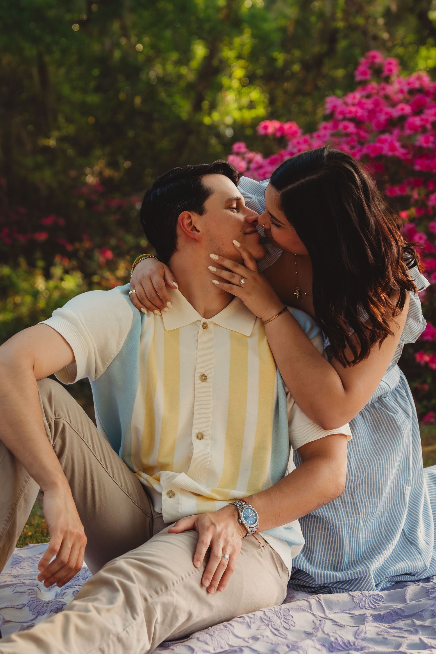 Couple sharing a romantic kiss outdoors on a blanket, surrounded by lush greenery and vibrant pink flowering bushes.