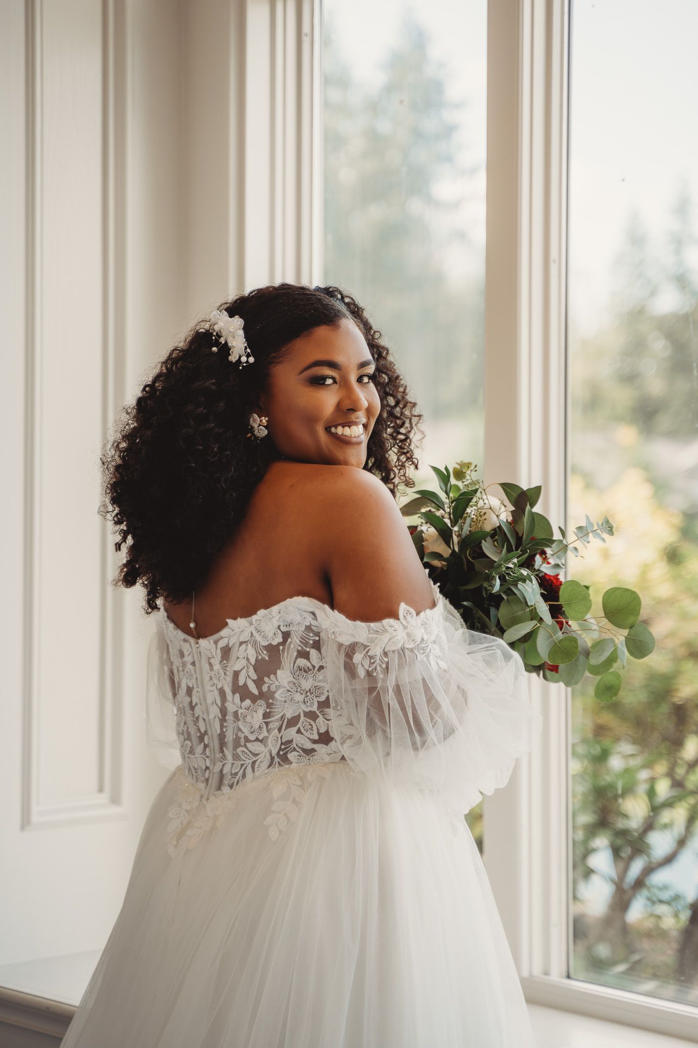 Smiling bride in off-shoulder lace wedding gown holds floral bouquet near window with curly hair and pearl accessories.