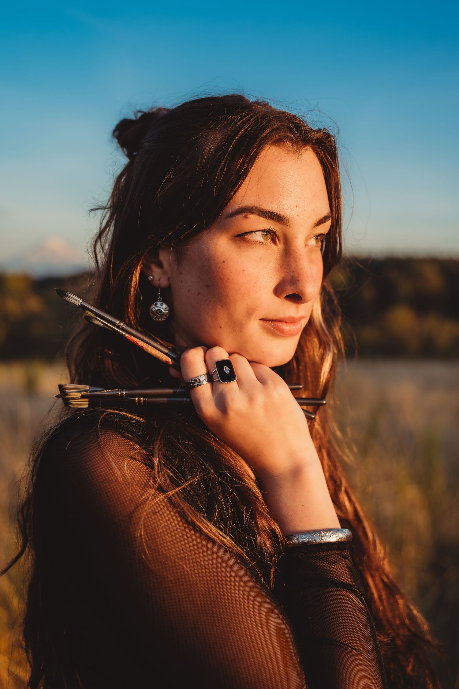 Young woman with auburn hair holds arrows over shoulder, wearing silver rings, in golden hour outdoor light.