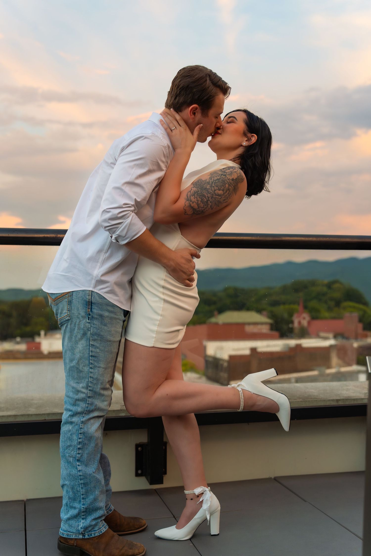Couple kissing on a rooftop terrace at sunset, she in a white dress and heels with one leg raised, overlooking a town and mountains.