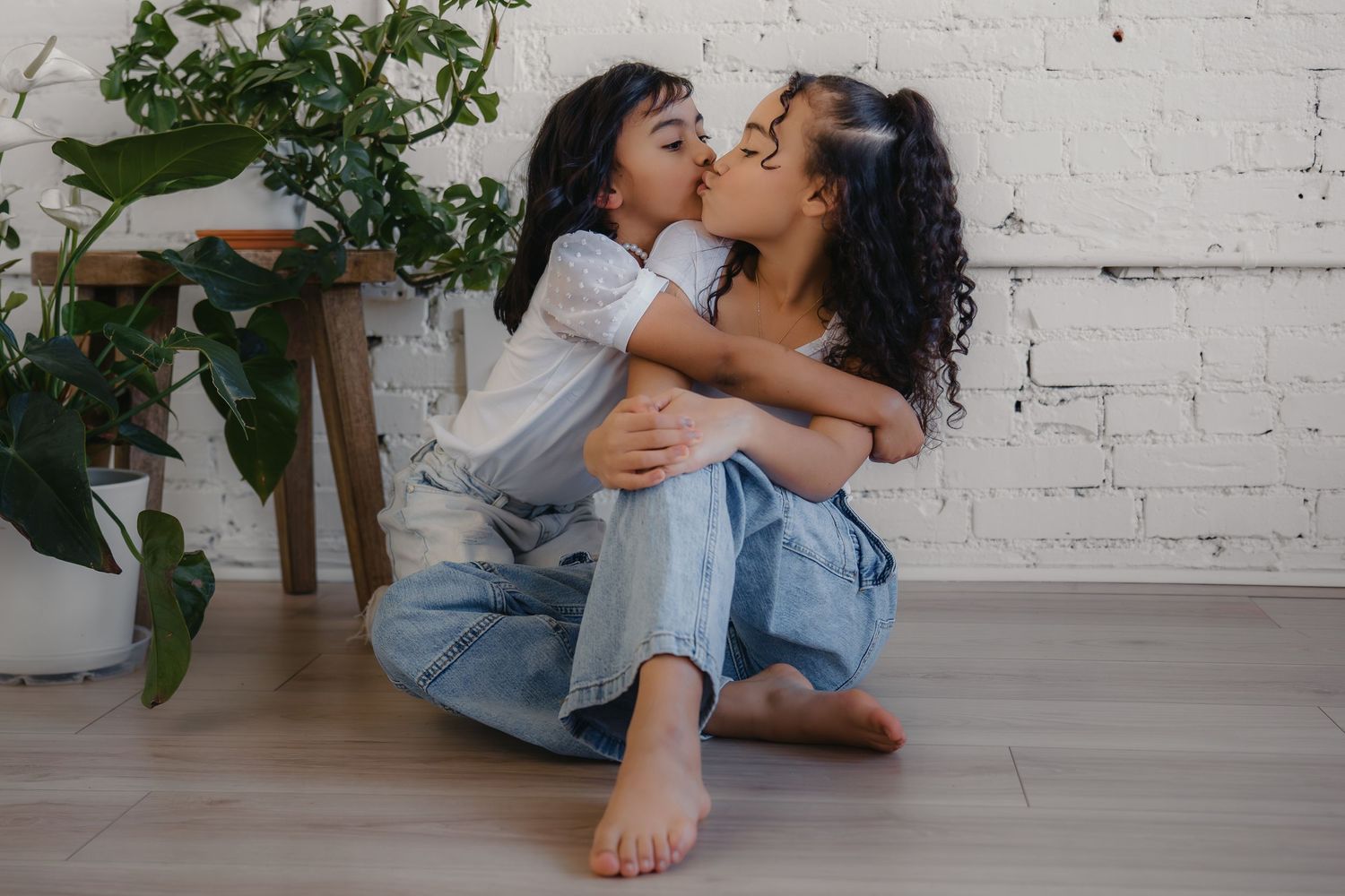 Two young girls sharing a sweet kiss while sitting on a hardwood floor near white brick wall and green plants.