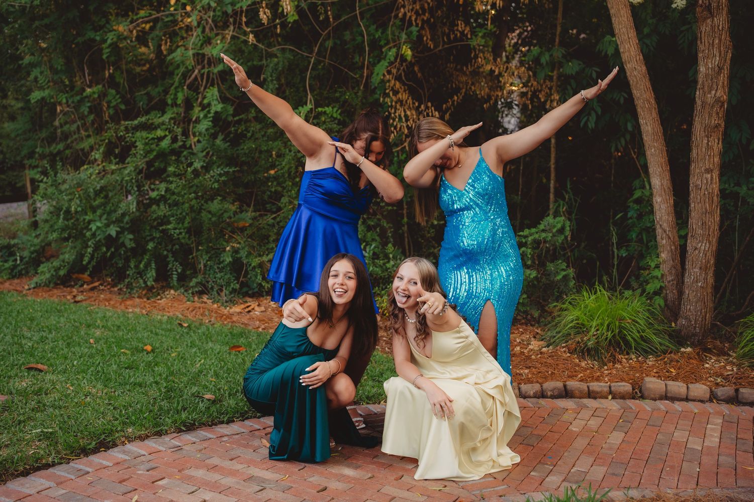Four young women in colorful formal dresses posing playfully outdoors on a brick path surrounded by lush green trees.