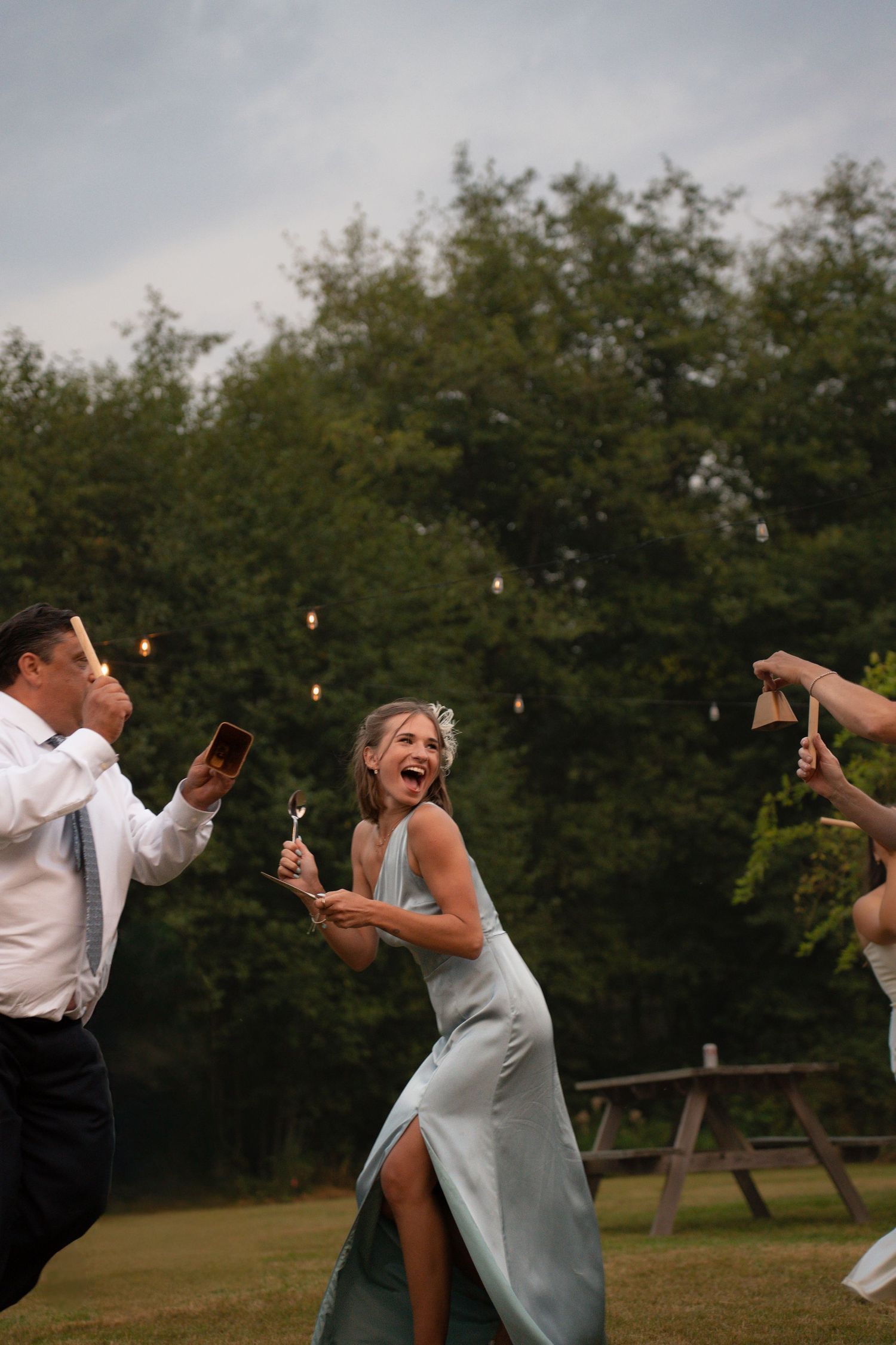 Woman in sage green gown laughing joyfully at outdoor wedding reception with guests holding cow bells at dusk.Charleston South Carolina