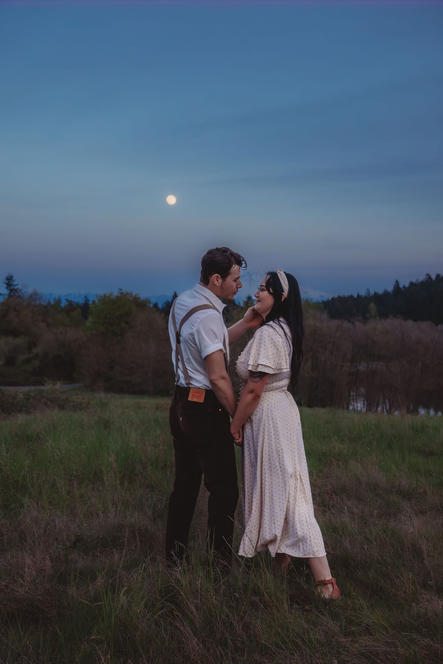 Romantic couple standing in a moonlit field at dusk, man in suspenders and woman in white dress holding hands.