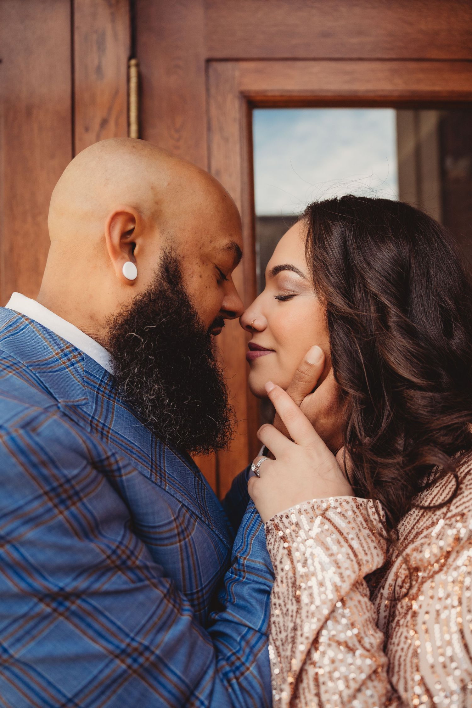 A bald bearded man in plaid suit tenderly holds woman's chin as they share an intimate moment, almost kissing.