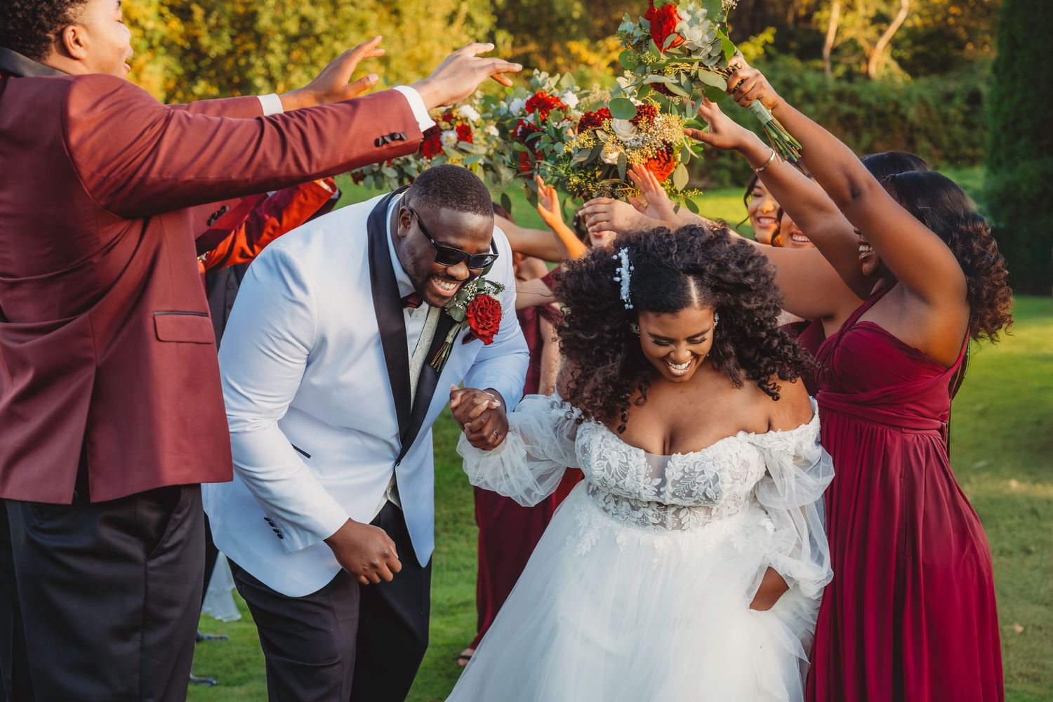 Joyful newlyweds walk under floral arch held by wedding party in red attire on lush green lawn.