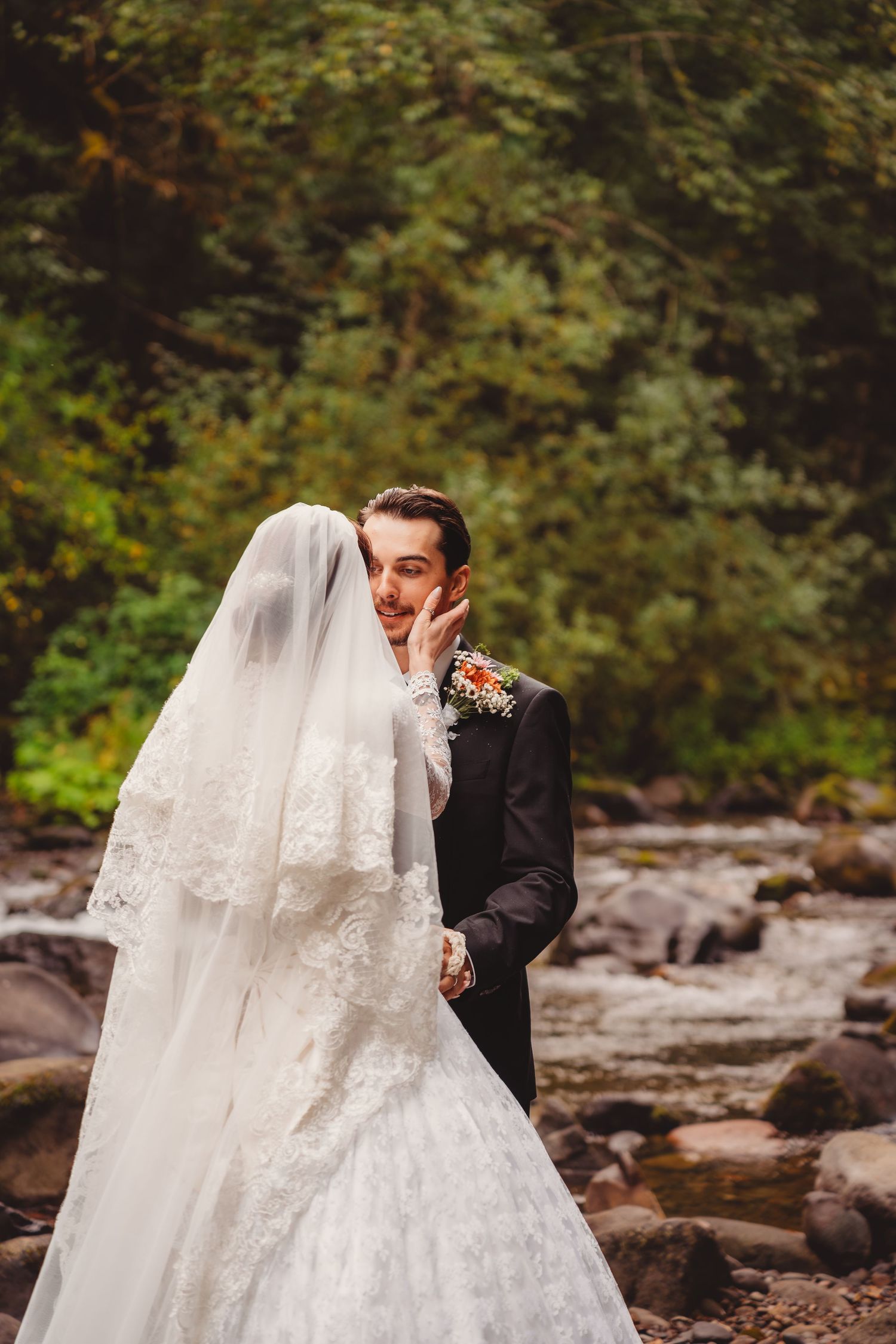 A bride in a white lace gown and veil embraces a groom in a black suit near a rocky river surrounded by lush greenery.