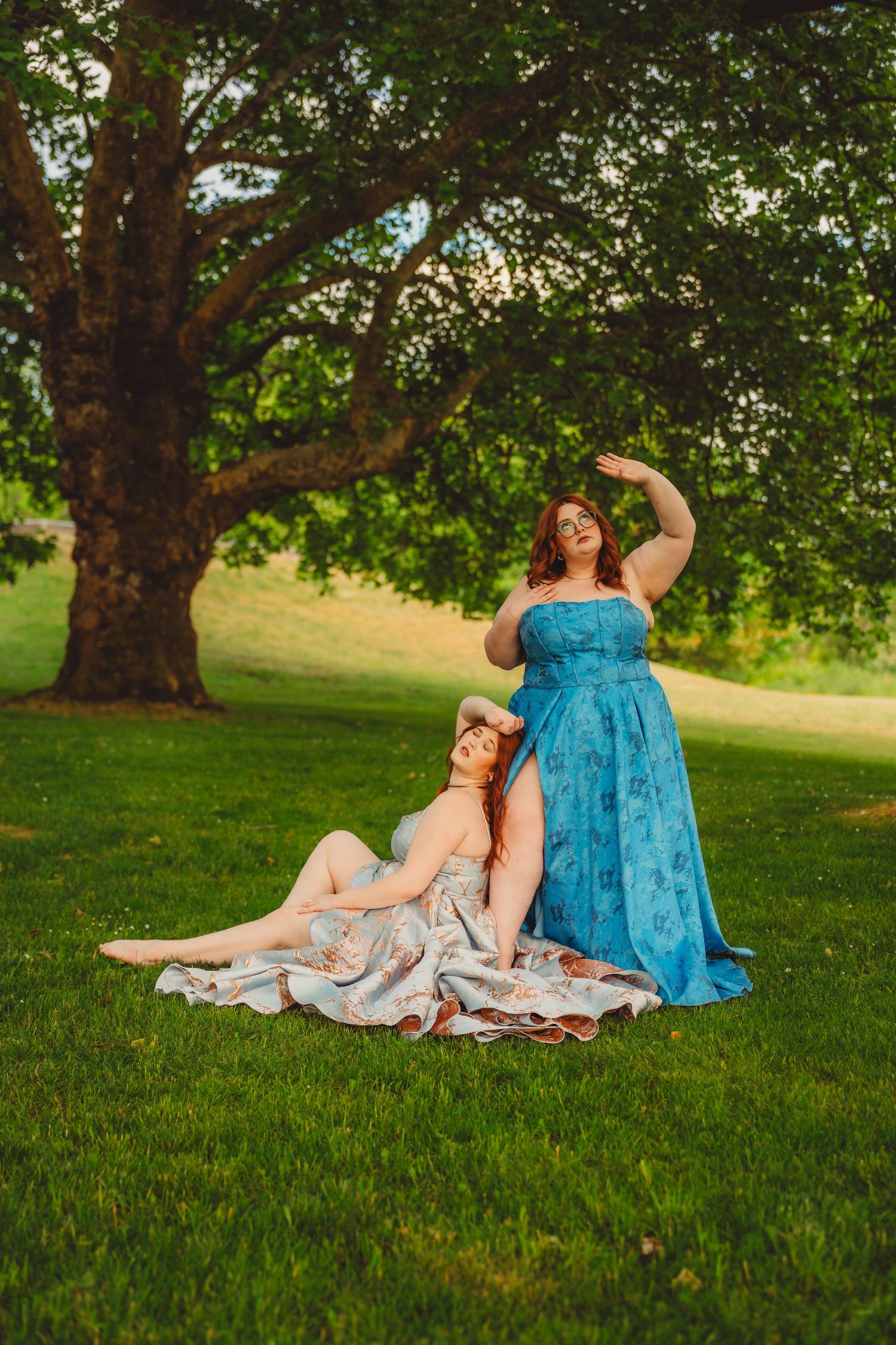 Two women in elegant dresses pose outdoors under a large tree on green grass in warm golden light.