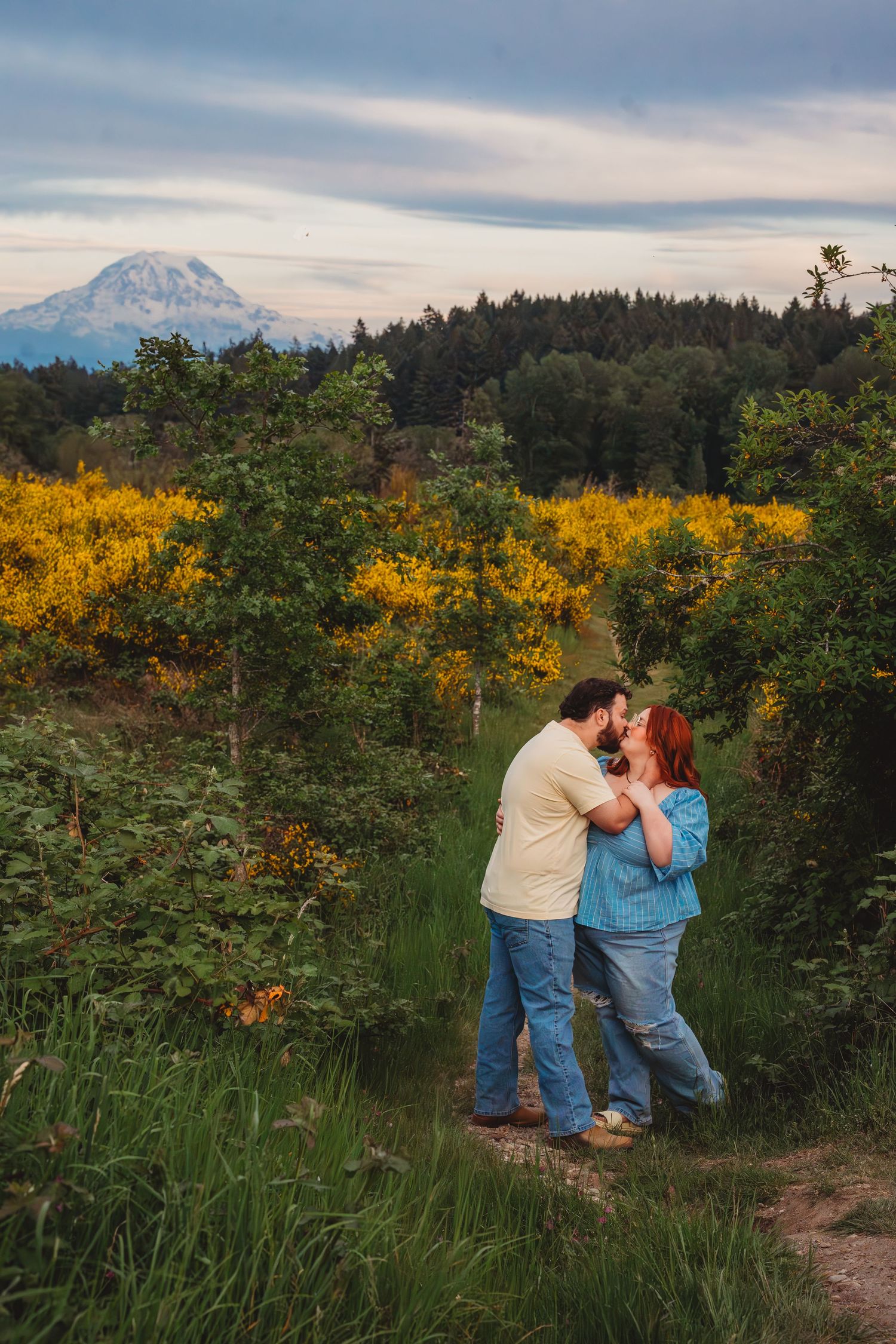Couple kissing on a trail surrounded by yellow wildflowers with Mount Rainier in the background at sunset.