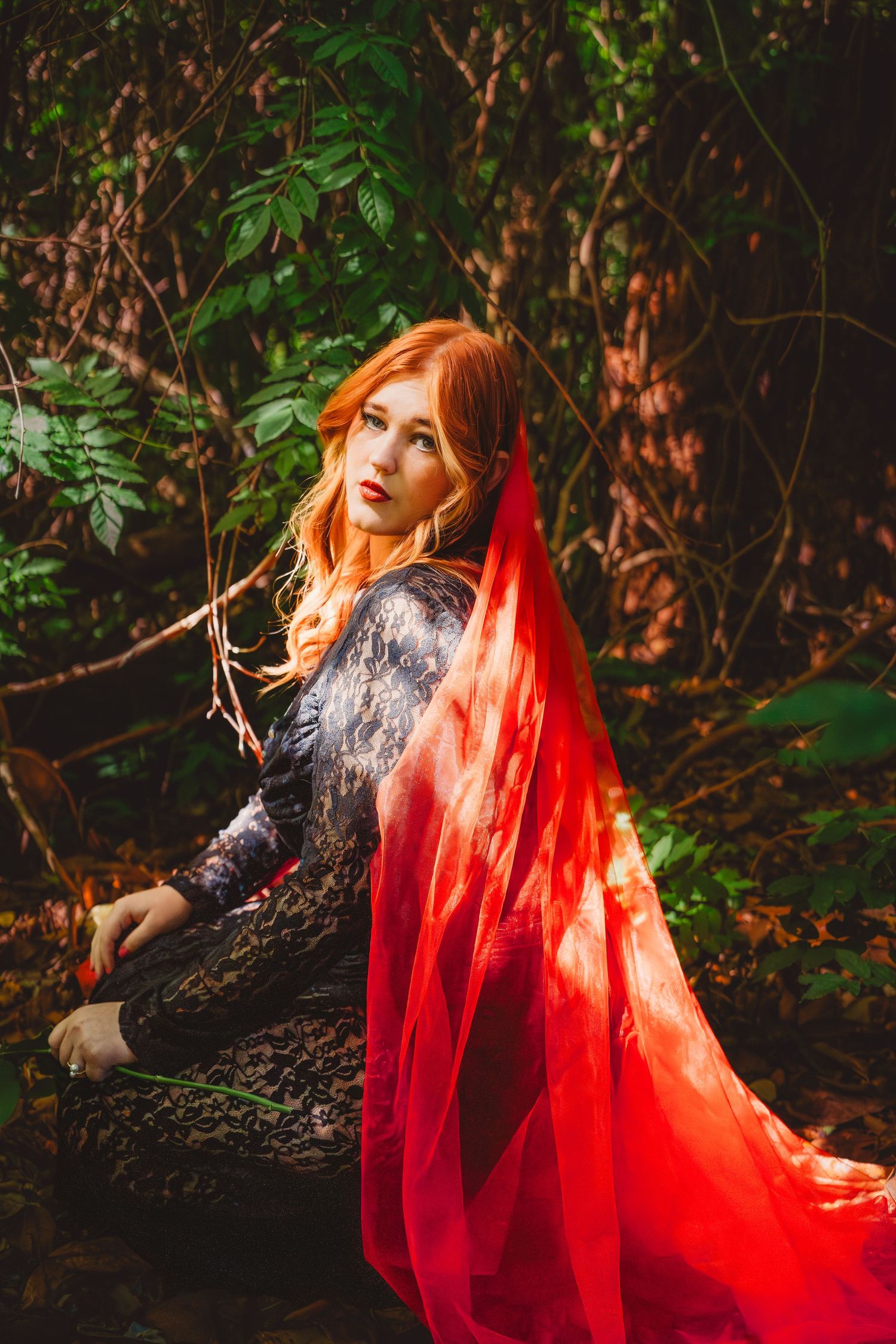 A red-haired woman in a lace dress and flowing red cape poses dramatically in a lush forest setting.