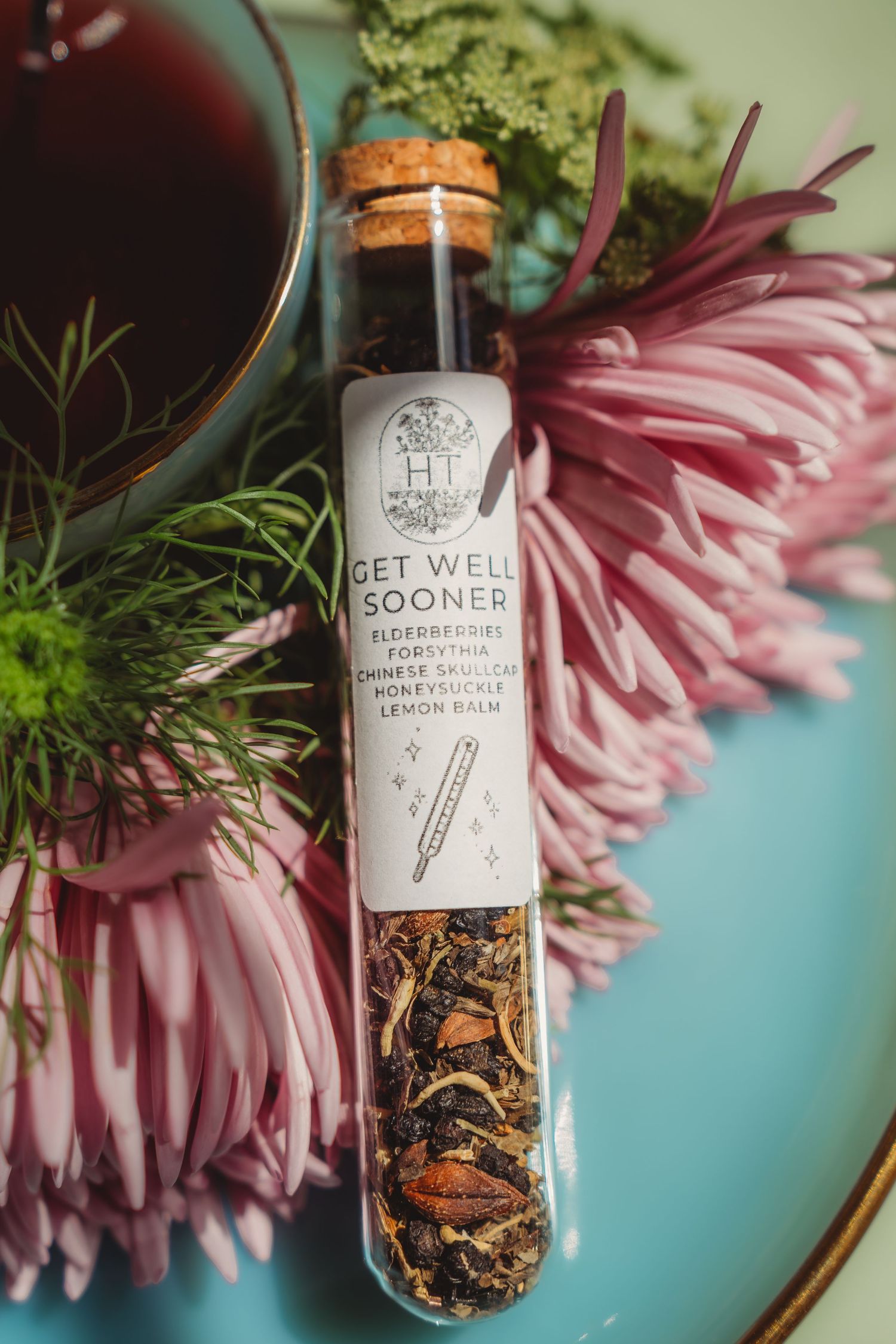 Glass tube with cork stopper containing Get Well Sooner herbal tea blend, surrounded by pink chrysanthemums on teal surface.