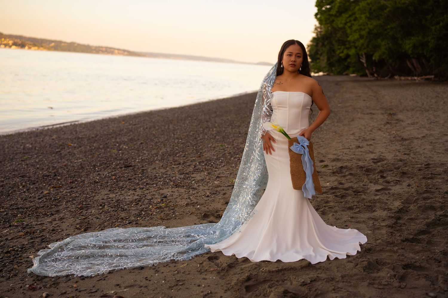Bride in white mermaid gown with lace veil on sandy beach at sunset.