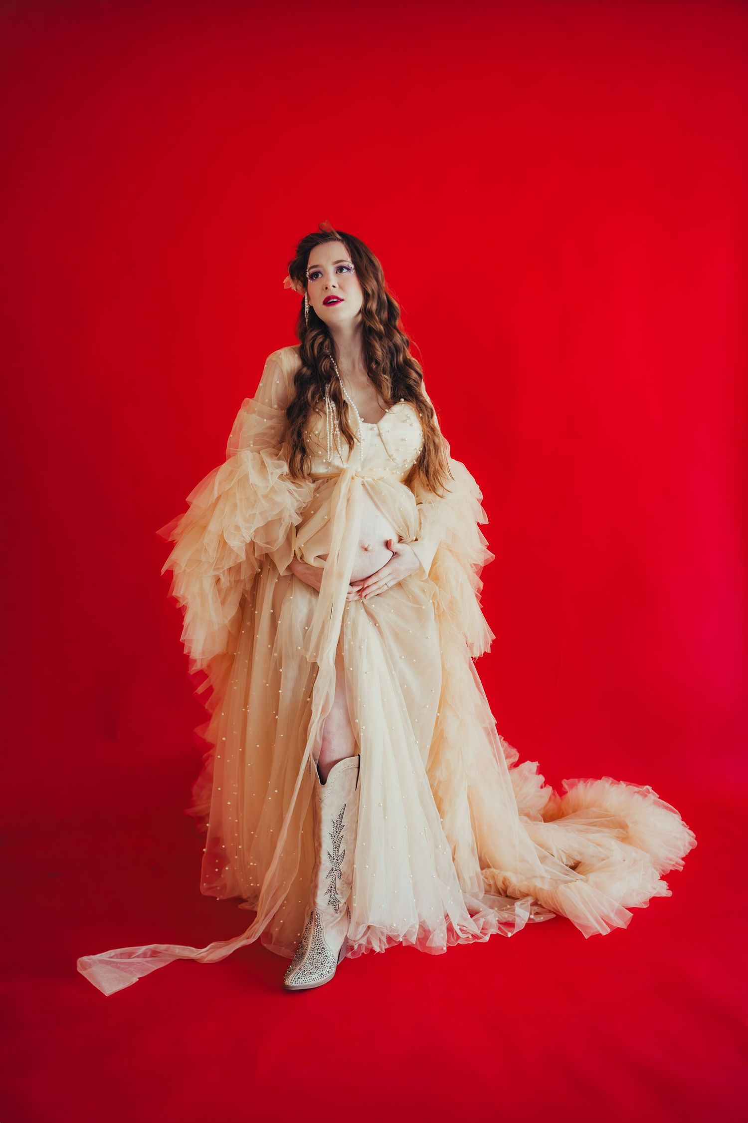 Pregnant woman in flowing cream tulle gown with ruffled sleeves poses against bold red backdrop in elegant maternity portrait.