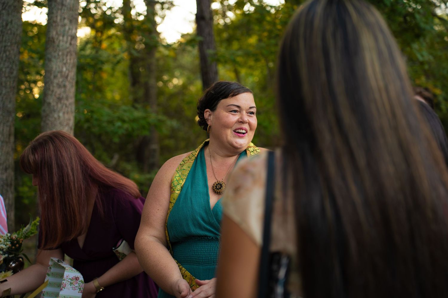 Smiling woman in a teal dress and gold-trimmed shawl laughs with friends gathered outdoors among trees in warm evening light.