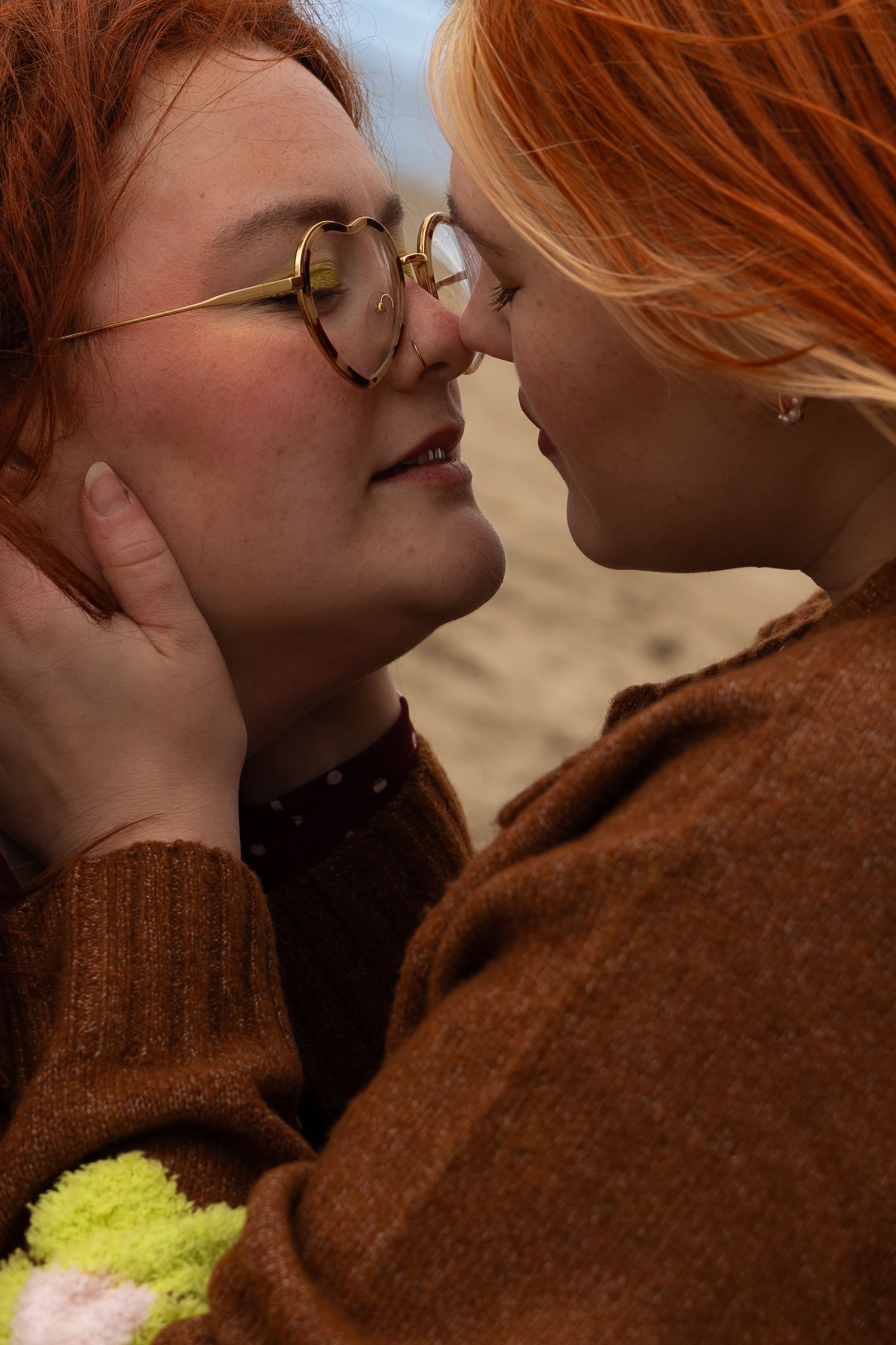 Two redhead women in brown sweaters share an intimate moment outdoors, nearly kissing, one wearing round gold glasses.
