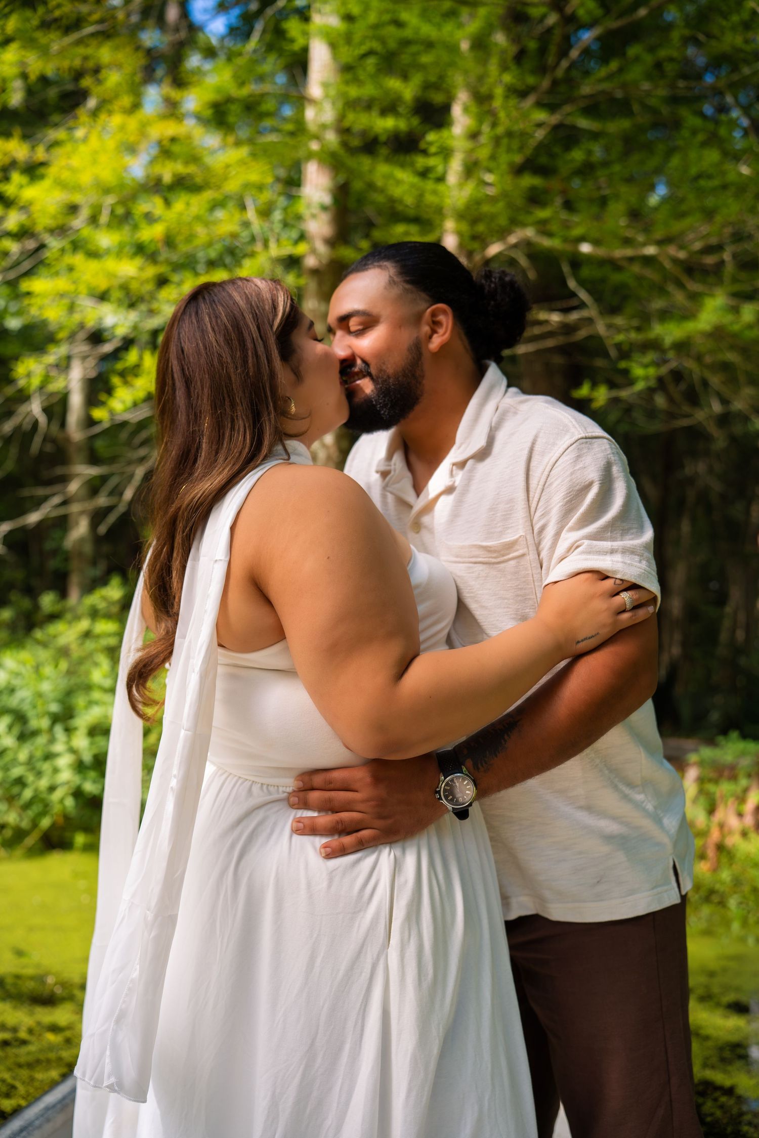 Couple in white outfits sharing a romantic kiss outdoors surrounded by lush green trees in a garden setting.
