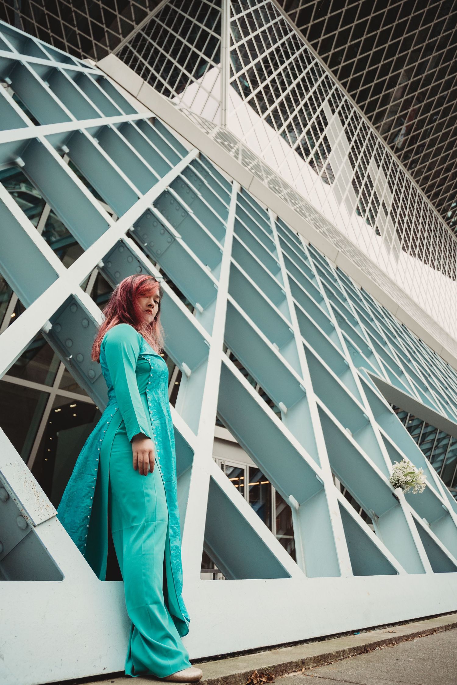 Woman with pink hair in teal jumpsuit poses against modern white geometric architectural structure.